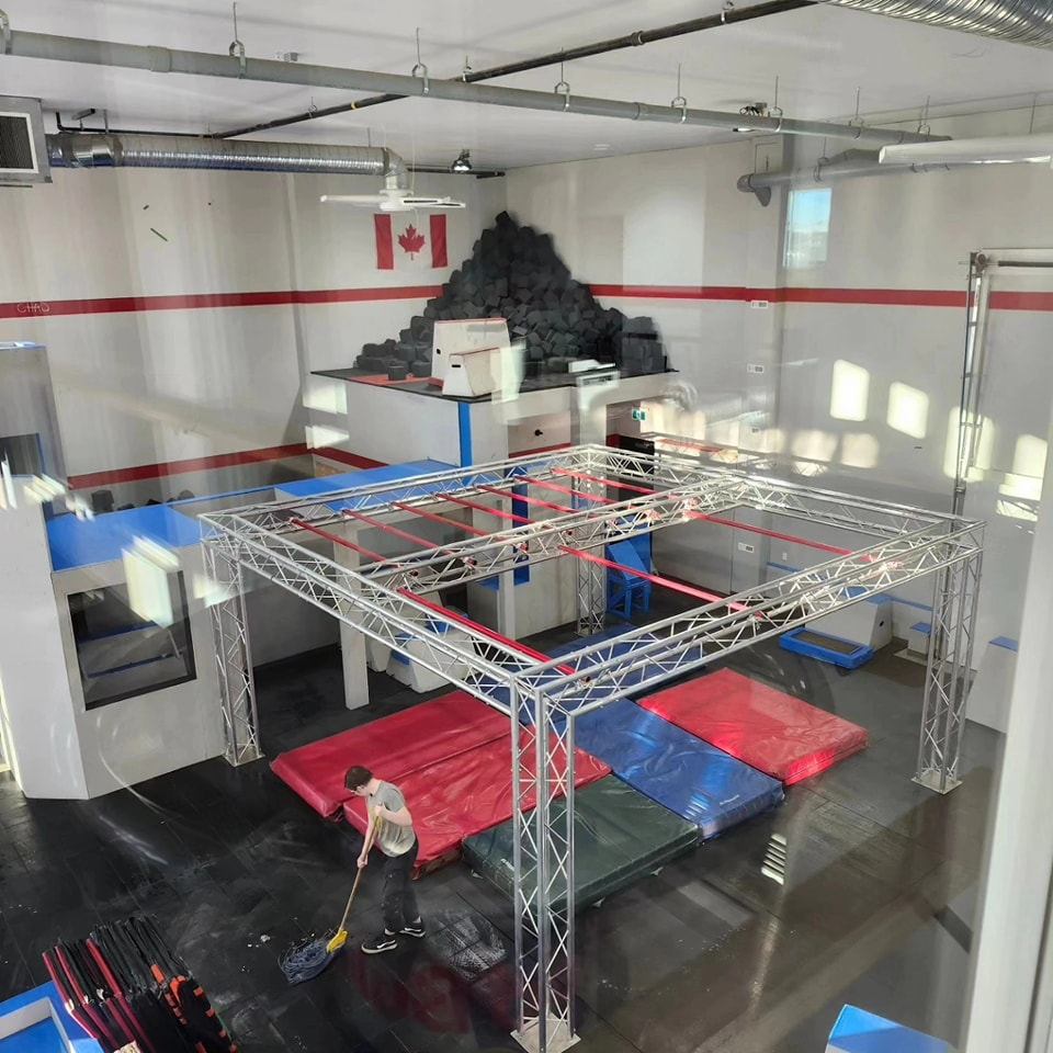 Indoor obstacle course gym with a person sweeping, mats, a climbing frame, and a Canadian flag on the wall.