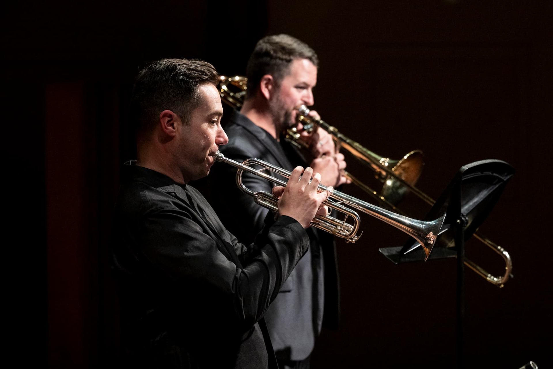 Brass musicians play trumpet and trombone on stage, facing music stands in darkness.