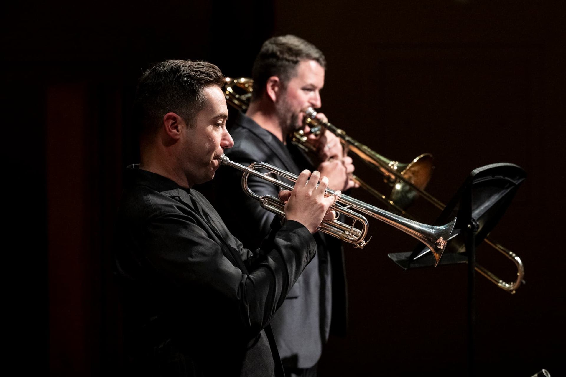 Brass musicians play trumpet and trombone on stage, facing music stands in darkness.