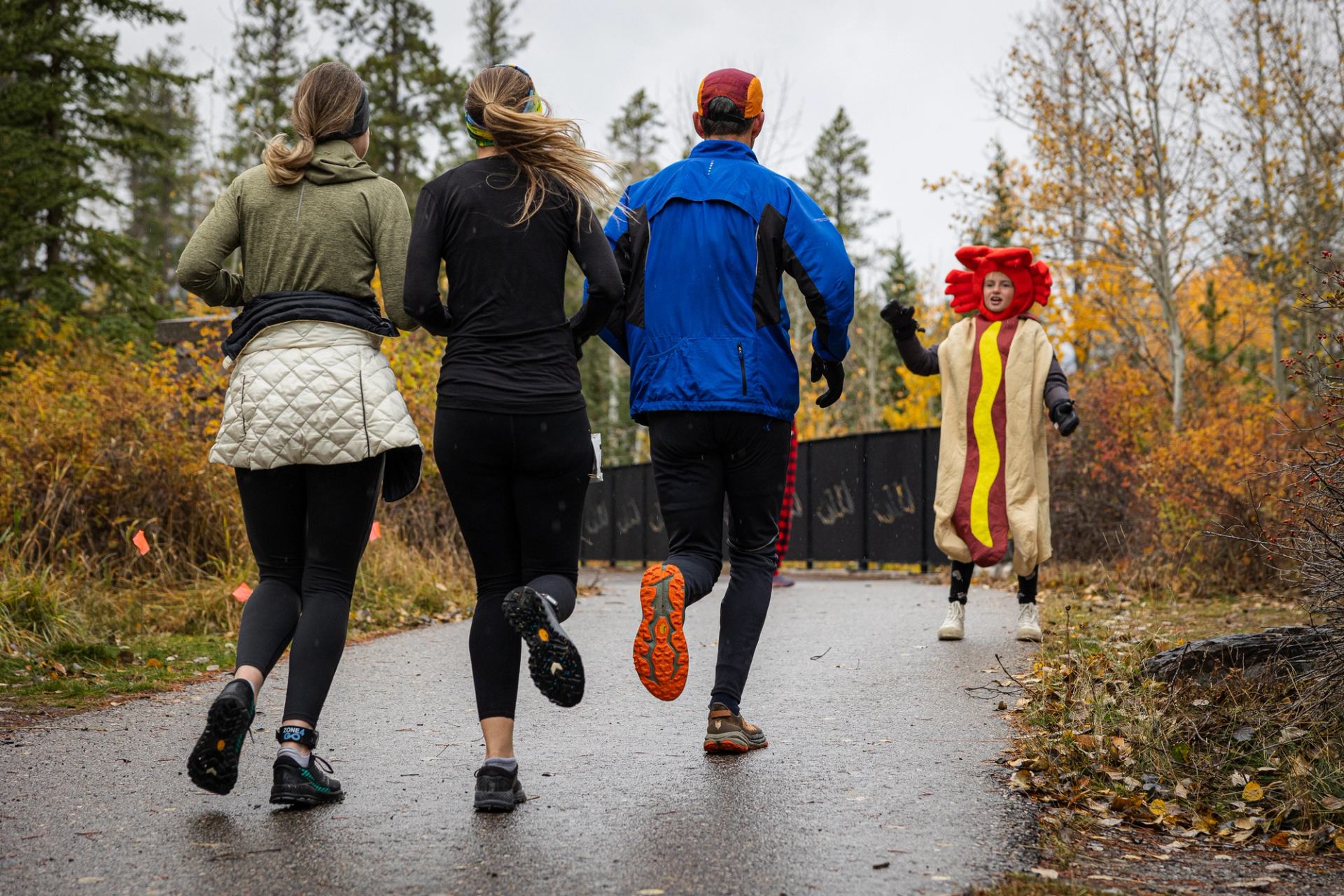 Runners on paved trail approach person in hot dog costume among autumn trees.