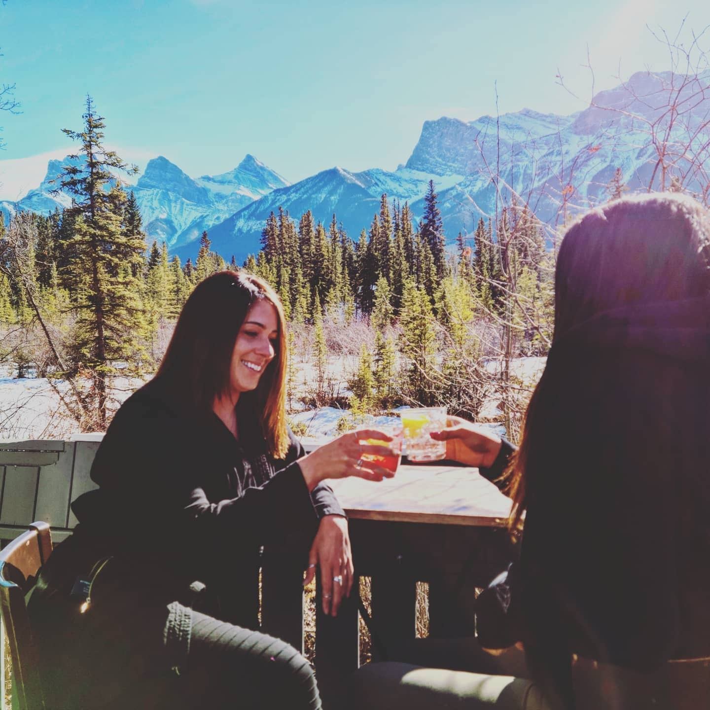 Two people toasting drinks outdoors with snowy mountains in the background.