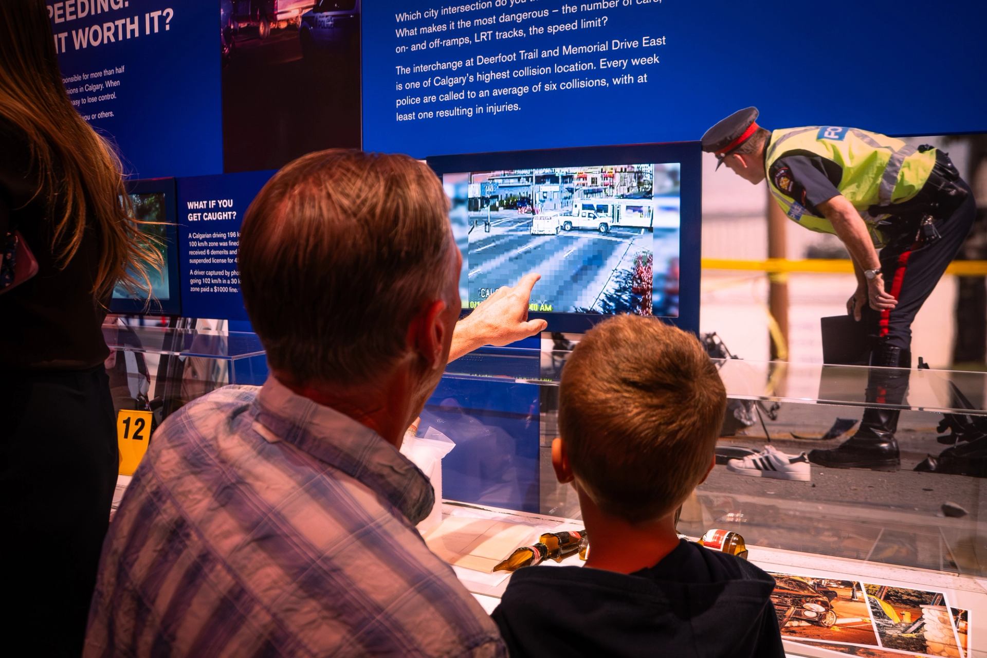 Museum visitors watch traffic collision footage on a screen as a police officer observes.