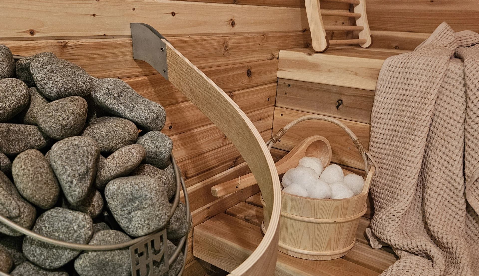 A close-up view inside a cedar wood sauna features a basket overflowing with smooth, volcanic sauna stones. Nearby, a small wooden bucket holds snowball-shaped ice pieces next to a folded waffle-weave towel.