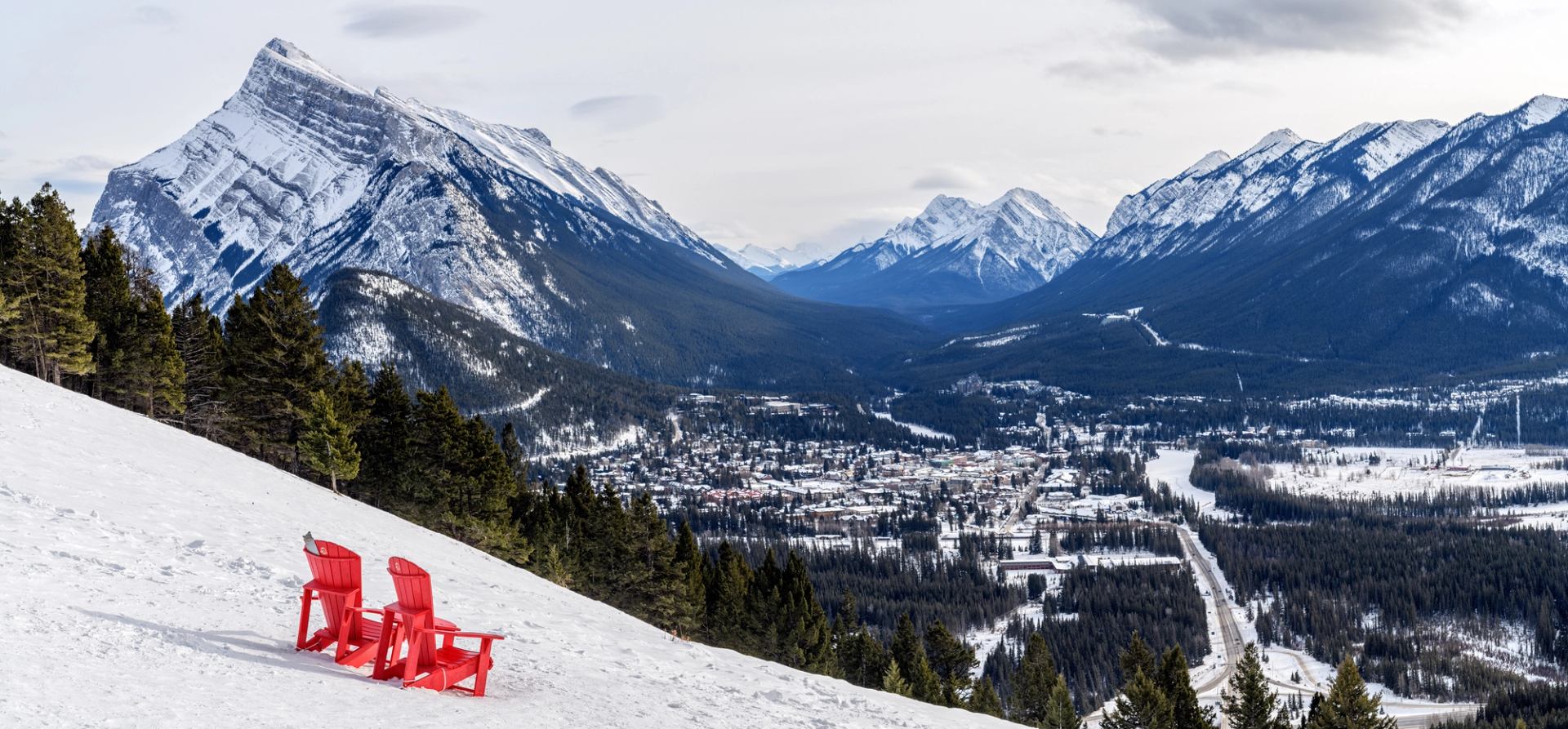 Two red Adirondack chairs on a snowy hill overlooking Banff valley and Rocky Mountains.