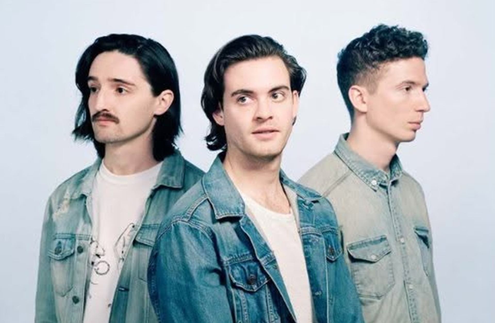 Three men in denim shirts stand in a row against a plain light background.