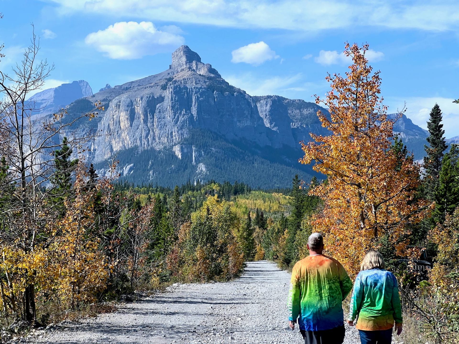 Two people walk along a gravel trail surrounded by autumn trees and towering mountains.