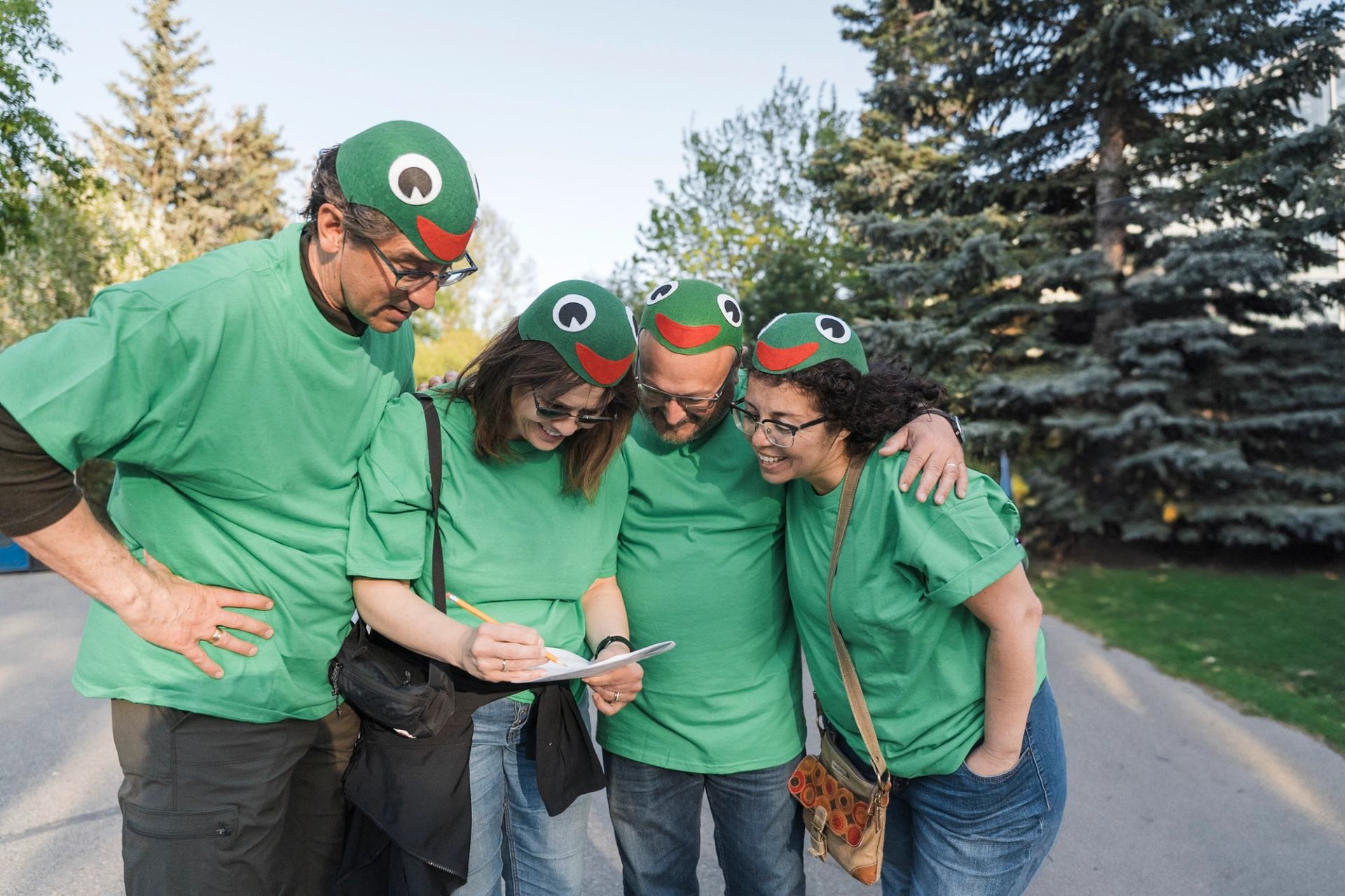 Group in matching green shirts and frog hats writes on clipboard in park.