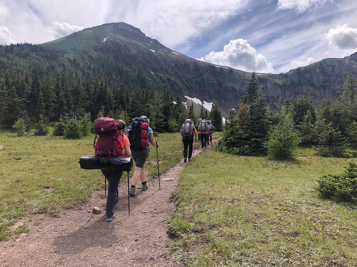 Hikers with large backpacks walk along a dirt trail through a mountain valley