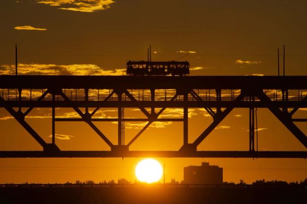 A city bus drives across a bridge with the sun setting in the background.