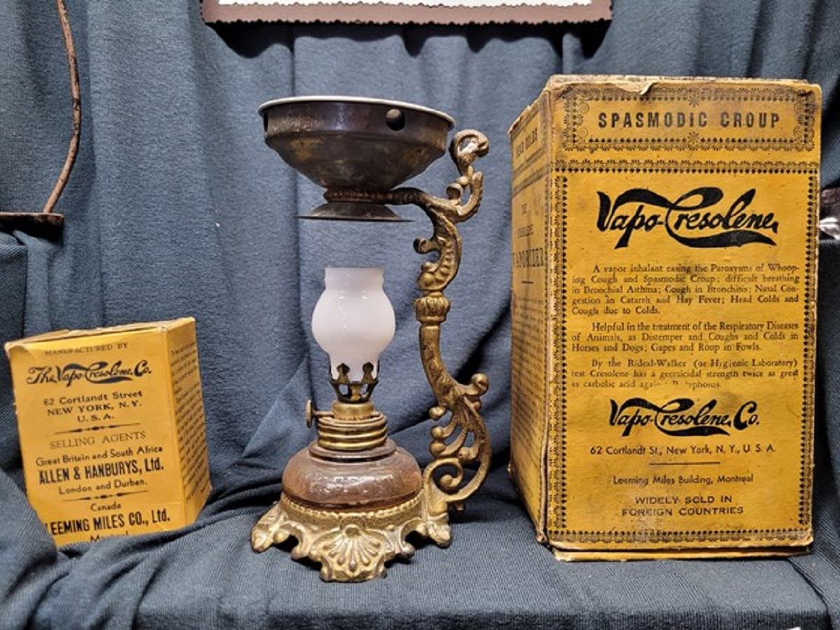 A vintage Vapo-Cresolene lamp beside its original yellow box, displayed on a dark fabric background. The lamp has a metal base and a small glass oil container.