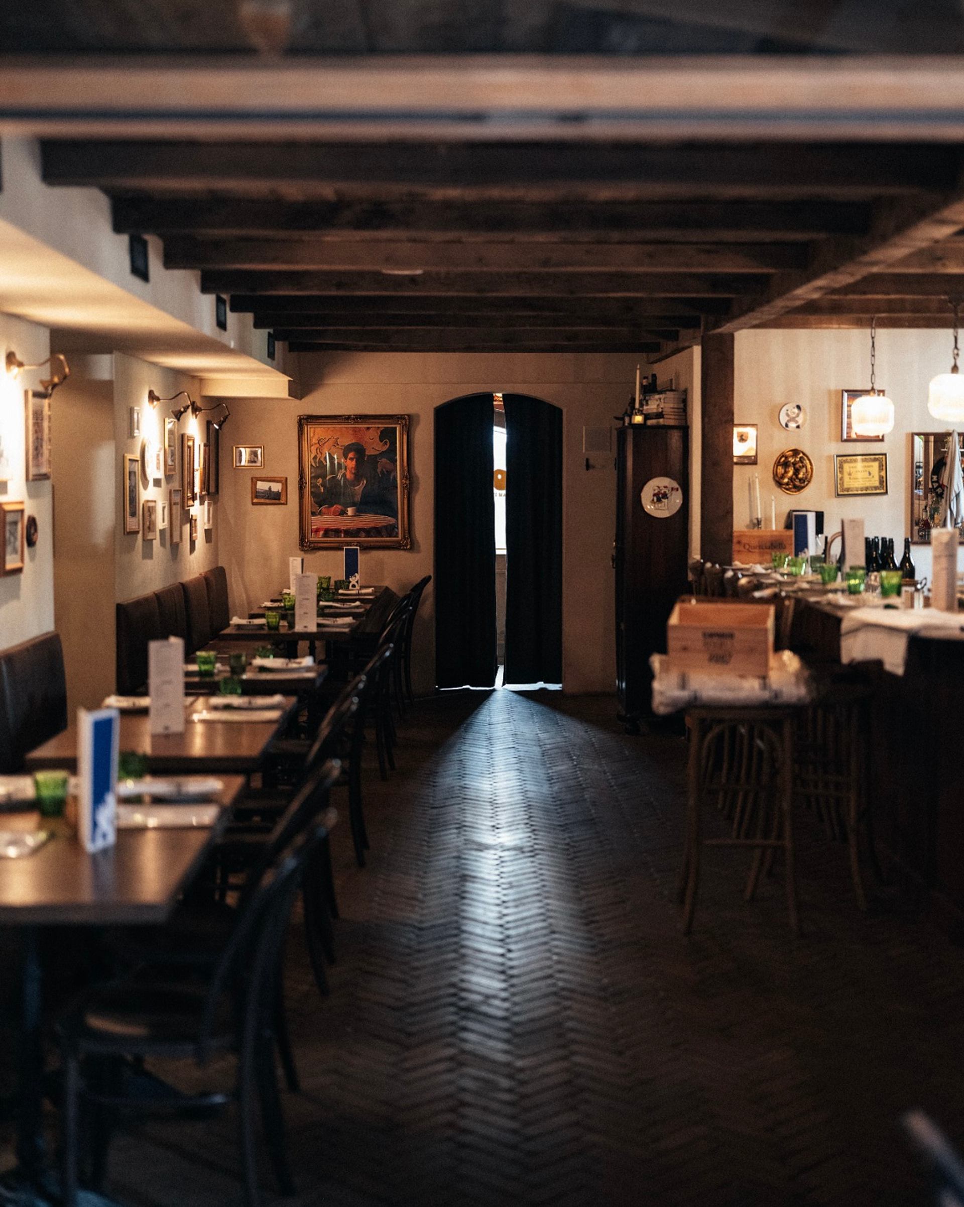 Narrow restaurant interior with set tables, bar counter, framed artwork and dark doorway.