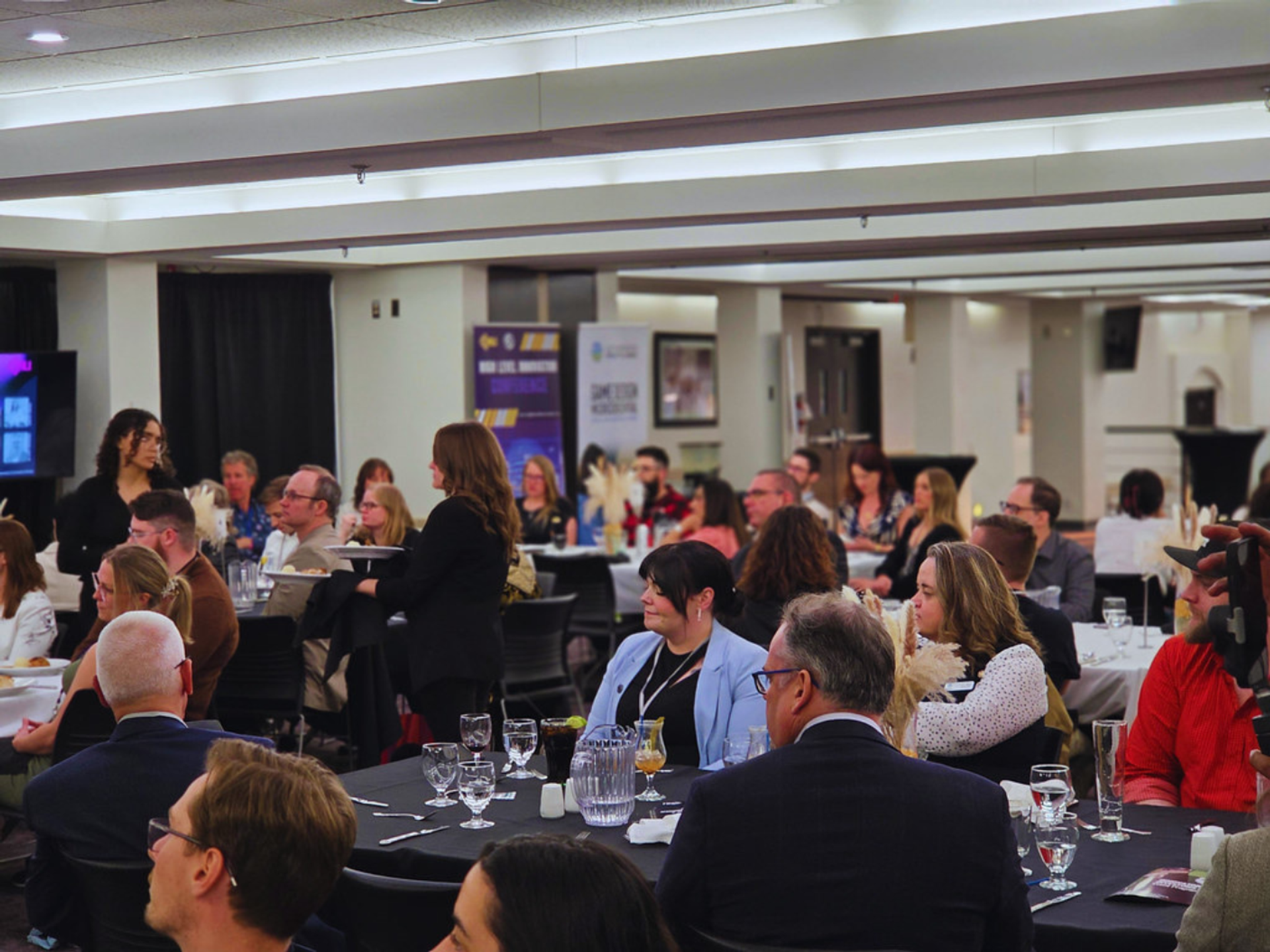 Large group seated at banquet tables during a conference event with a presenter speaking.