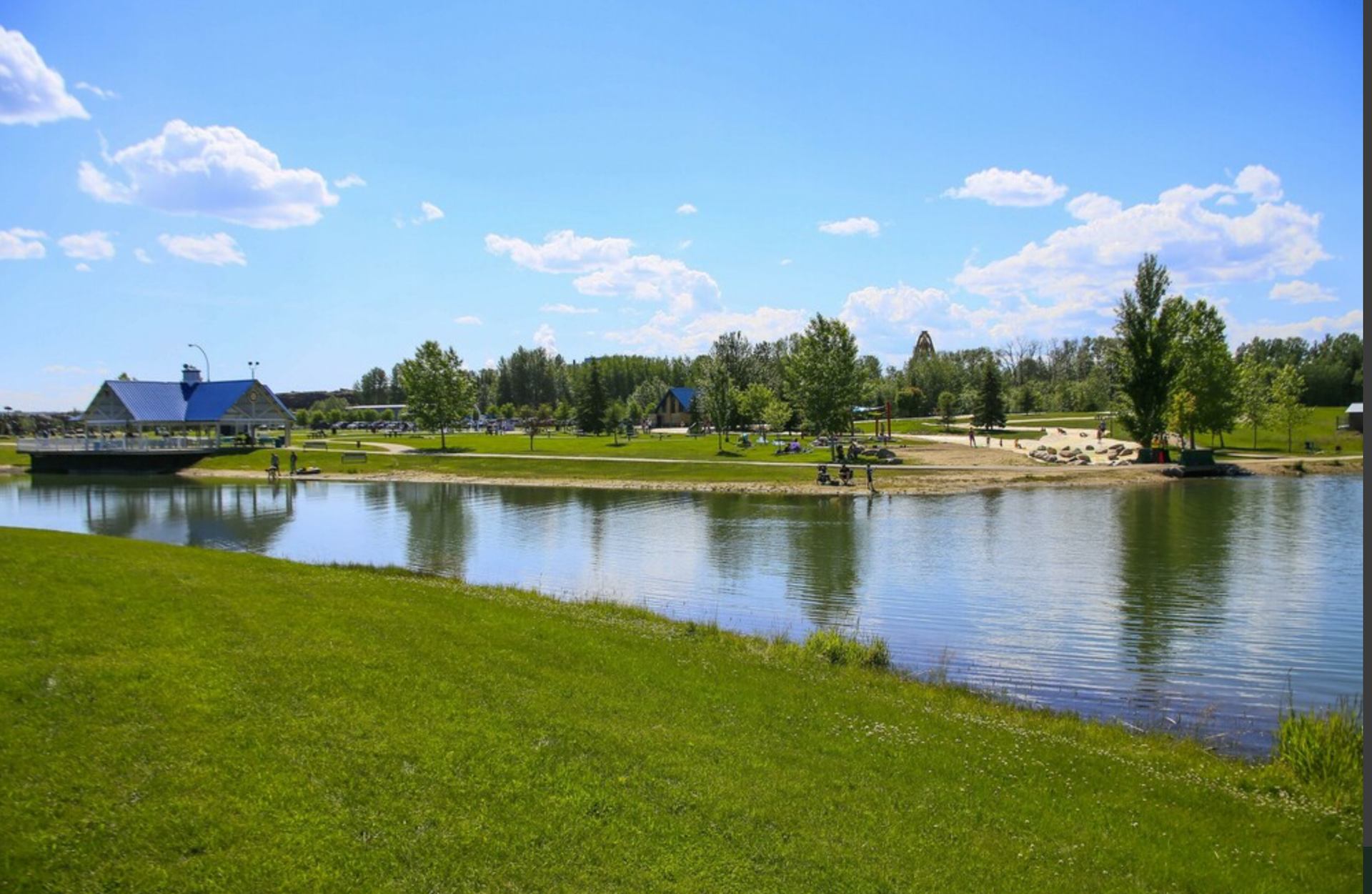 Festival Park scene featuring a calm pond bordered by lush green grass and sandy shores. Families are relaxing near the water, and a pavilion with a blue roof and trees provide shade in the background. The sky is blue with scattered white clouds.