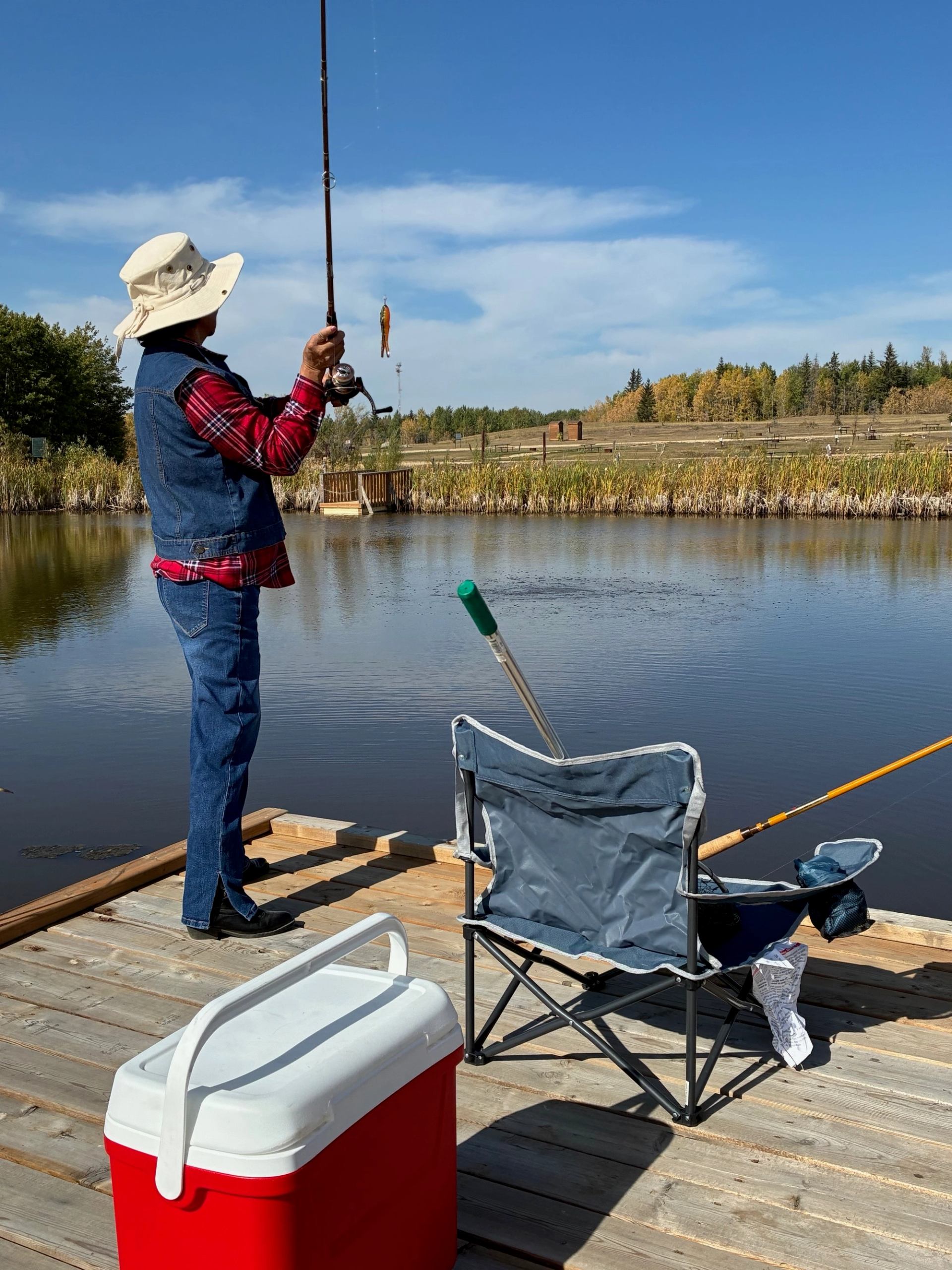 Person fishing from wooden dock beside folding chair, cooler and calm pond.