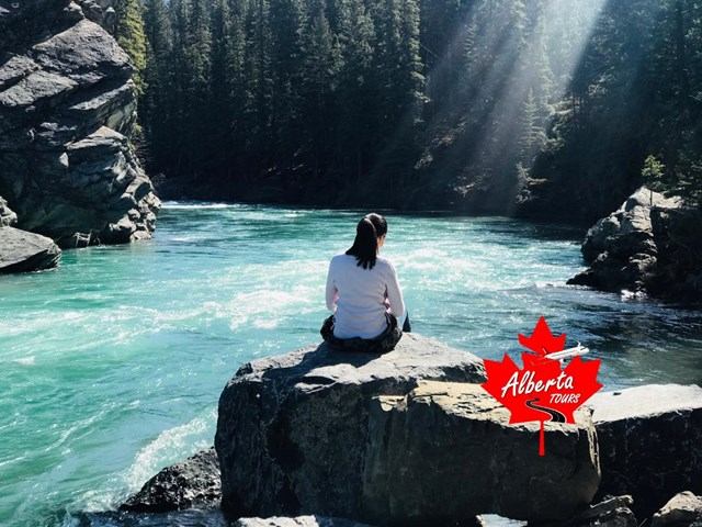 Person sitting on boulder beside turquoise river, forest canyon, Alberta Tours logo.