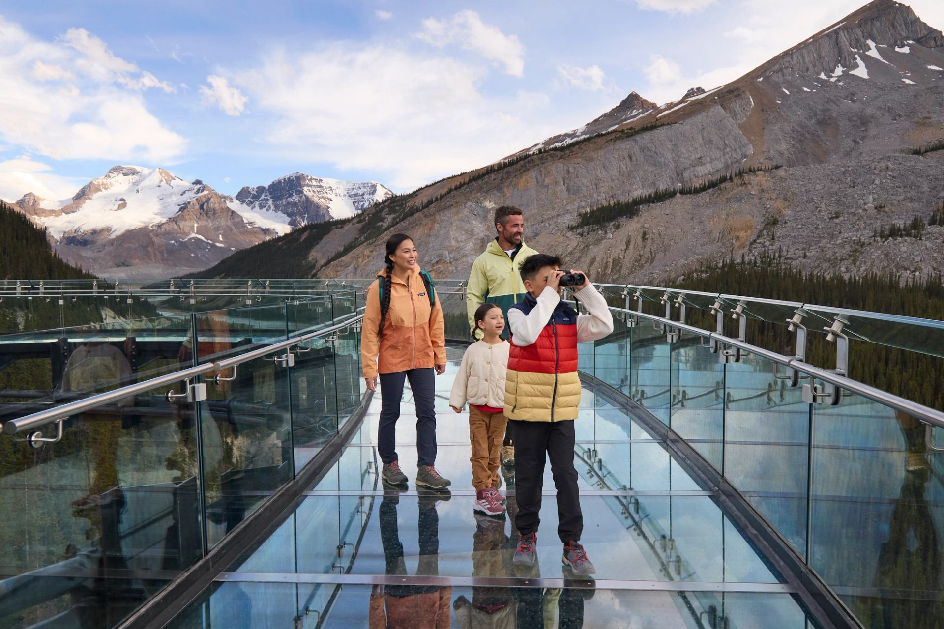 People walking on a glass bridge, with binoculars, overlooking snowy mountains and forest.