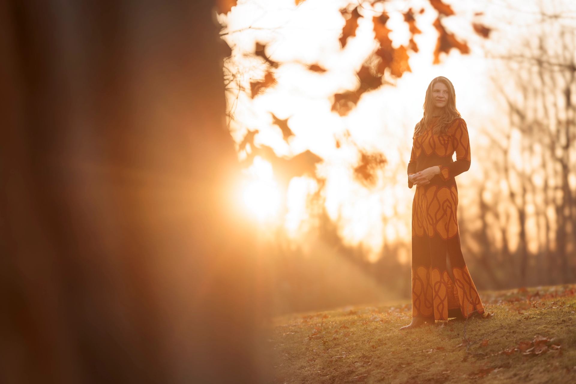 Person in long patterned dress stands on grassy slope with bright sun behind.