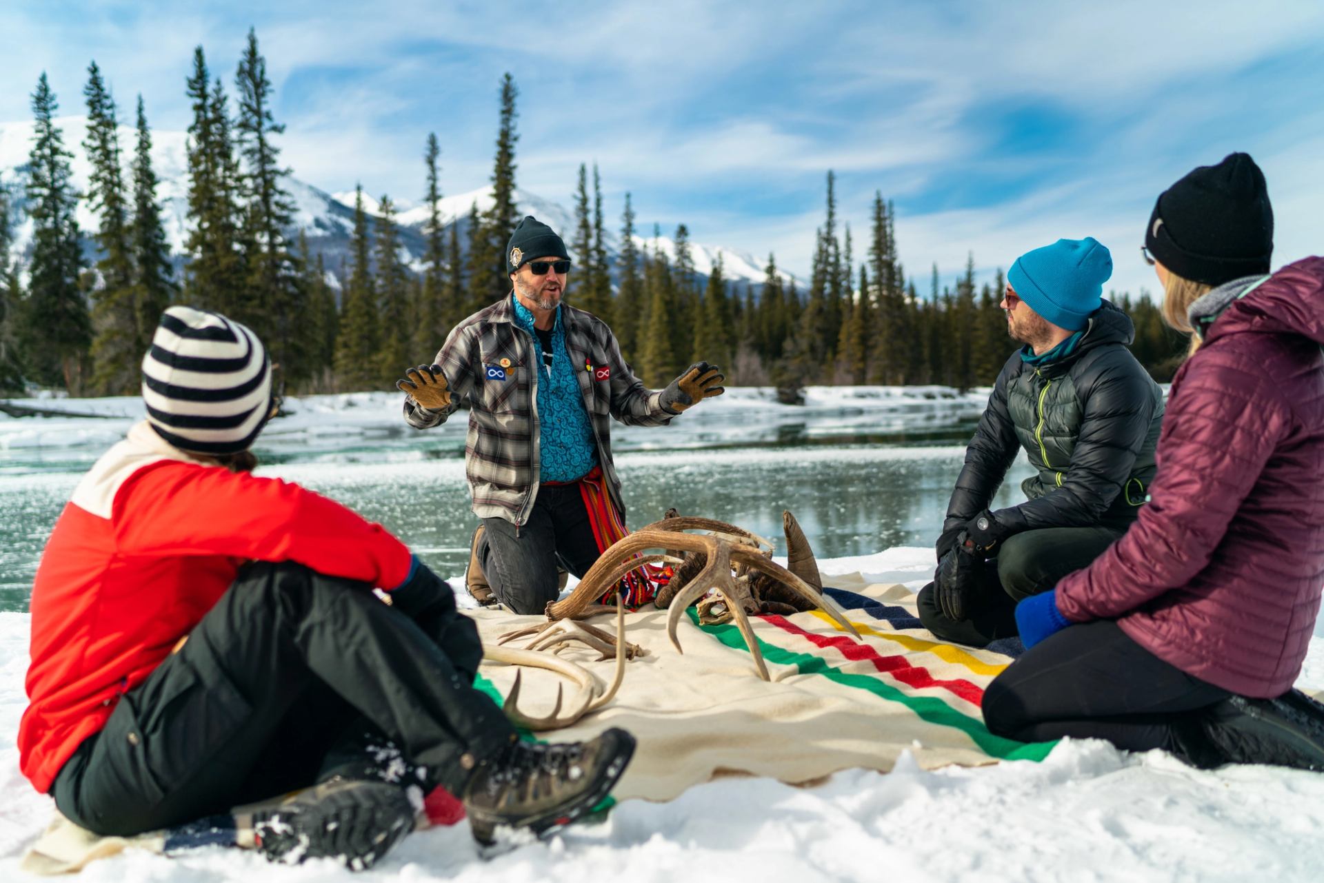 Four people sit on snowy riverbank listening, antlers on colorful blanket; evergreens and mountains behind.