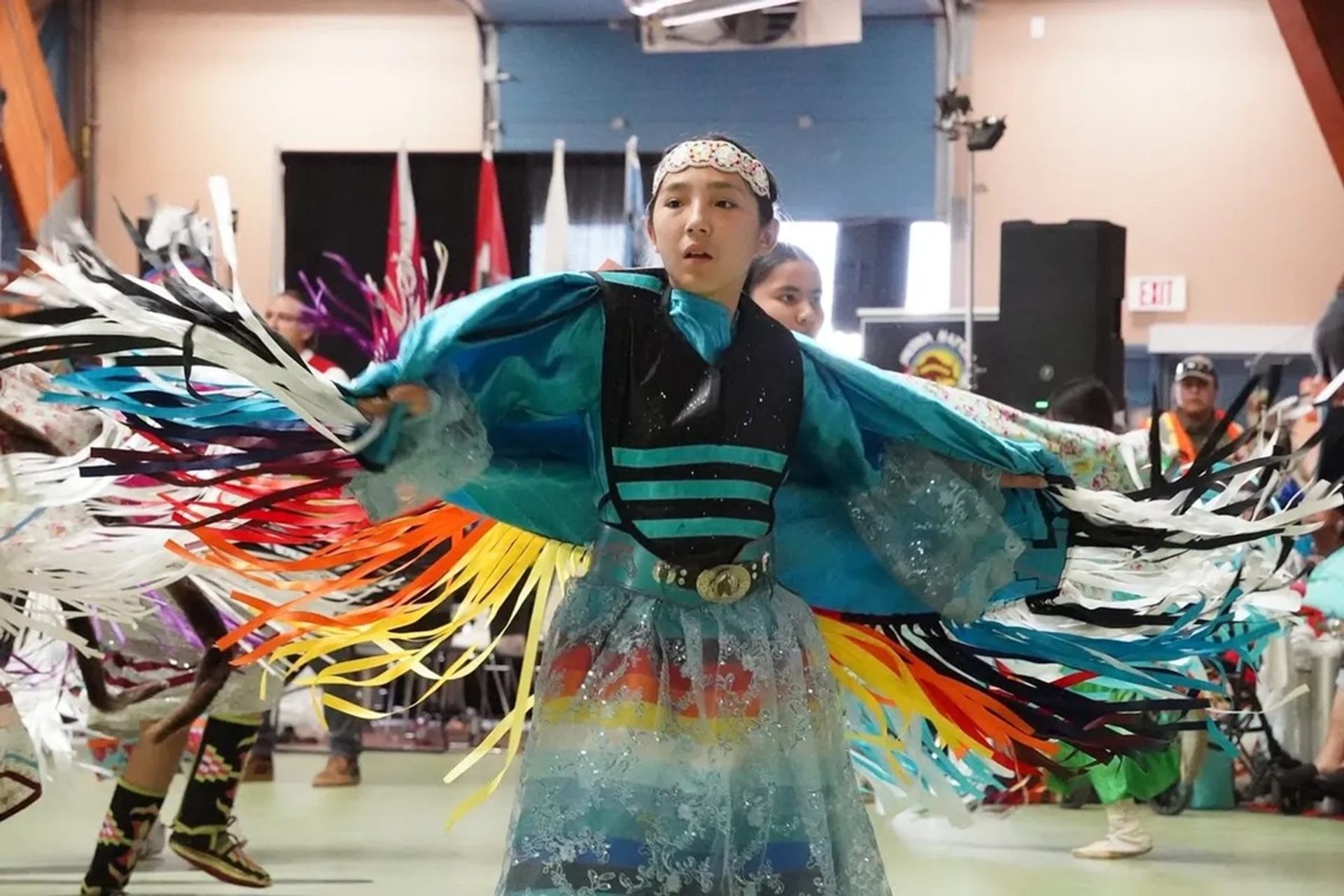 Powwow dancer in turquoise regalia spins with colourful fringe inside a community hall.