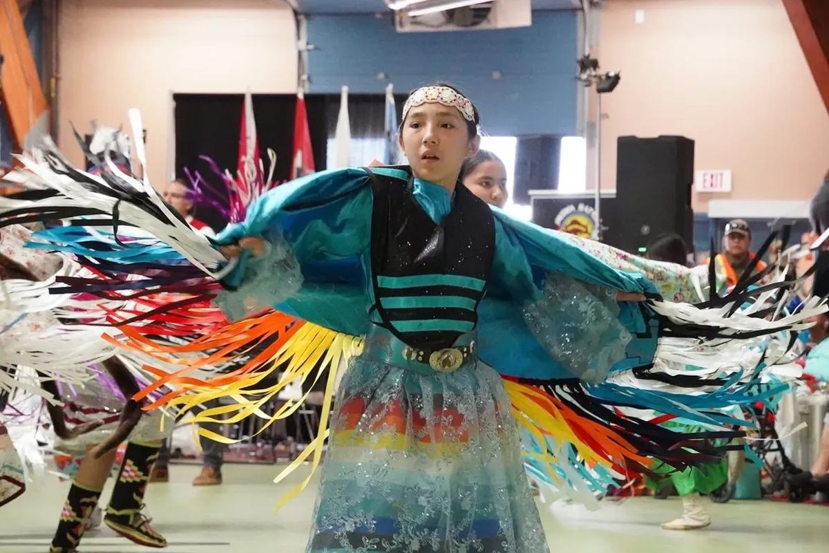 Powwow dancer in turquoise regalia spins with colourful fringe inside a community hall.
