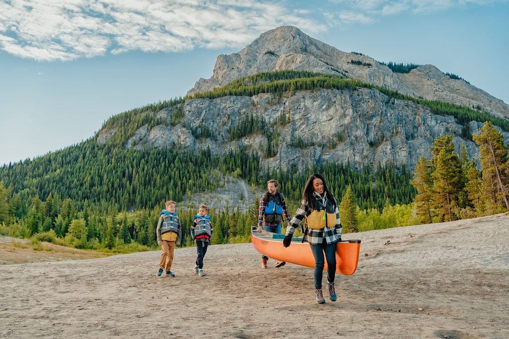 Four people carry an orange canoe across sand with forested mountains in the background.