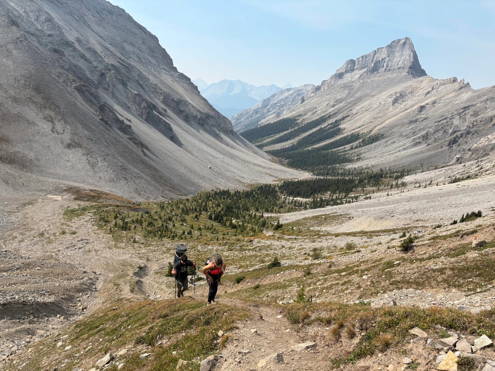Two hikers on a rocky alpine trail surrounded by towering gray mountains.