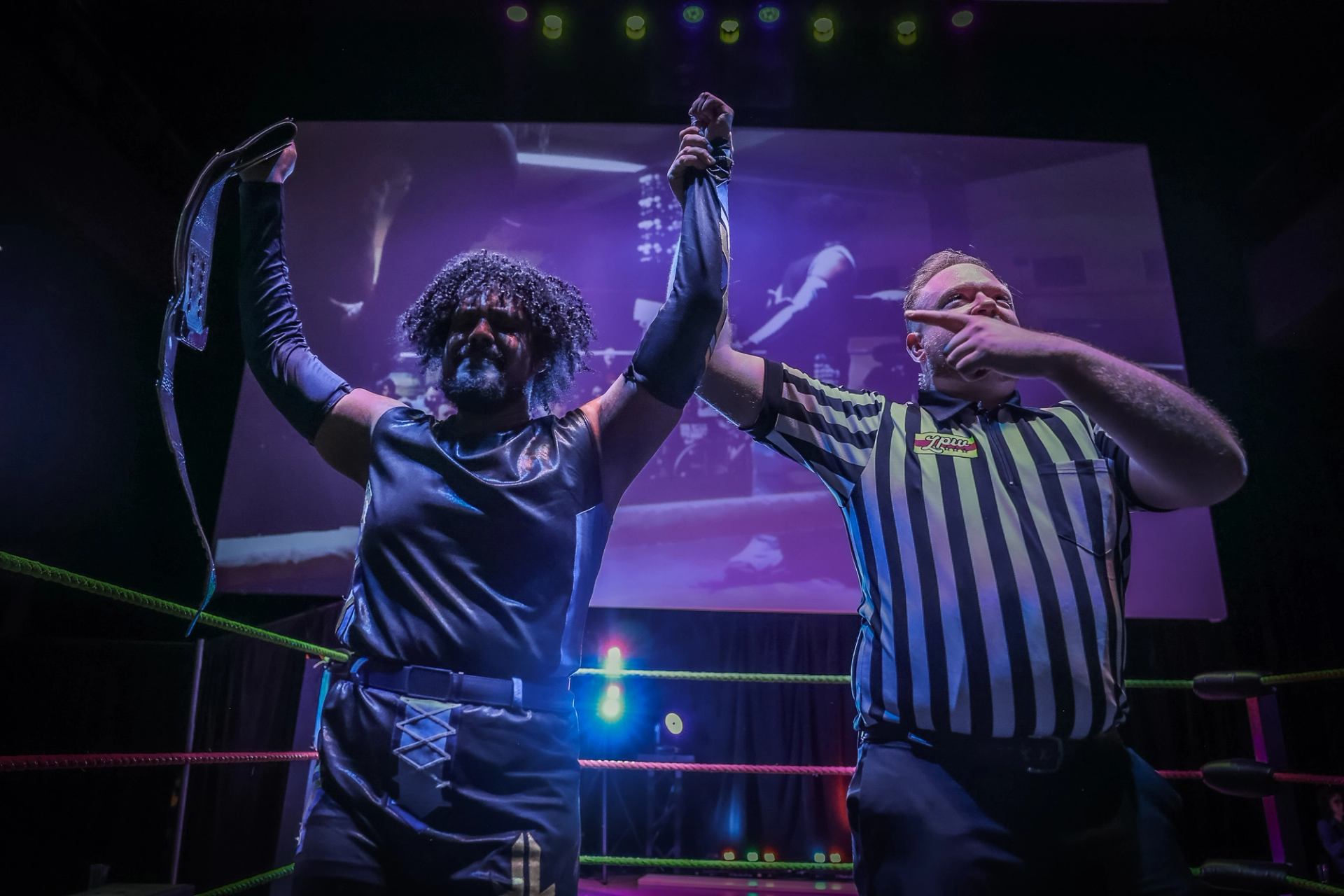 Wrestler raises championship belt with referee holding arm under stage lights.