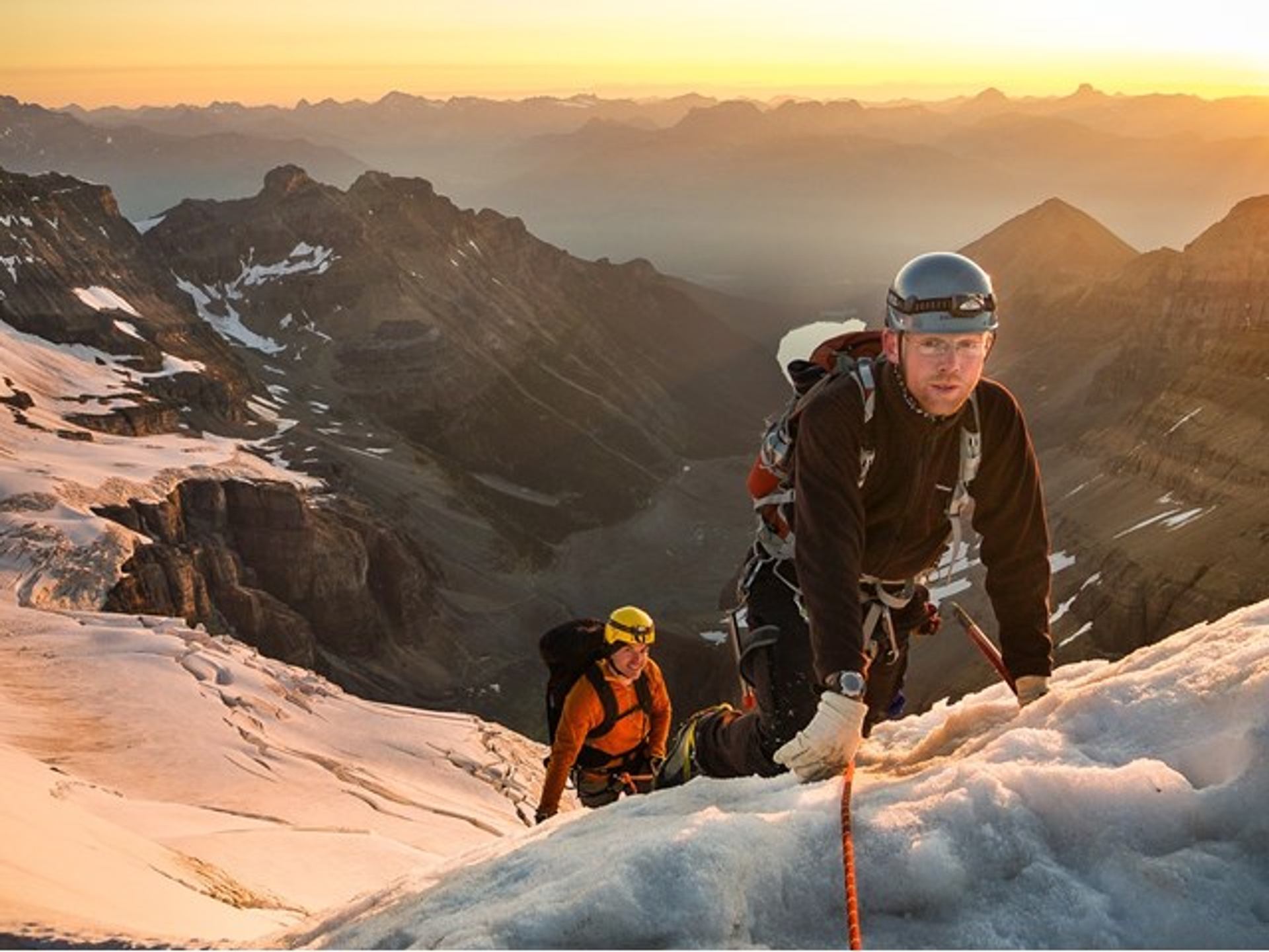 Group of climbers roped together ascending a snowy alpine ridge at sunrise with rugged mountain peaks beyond.
