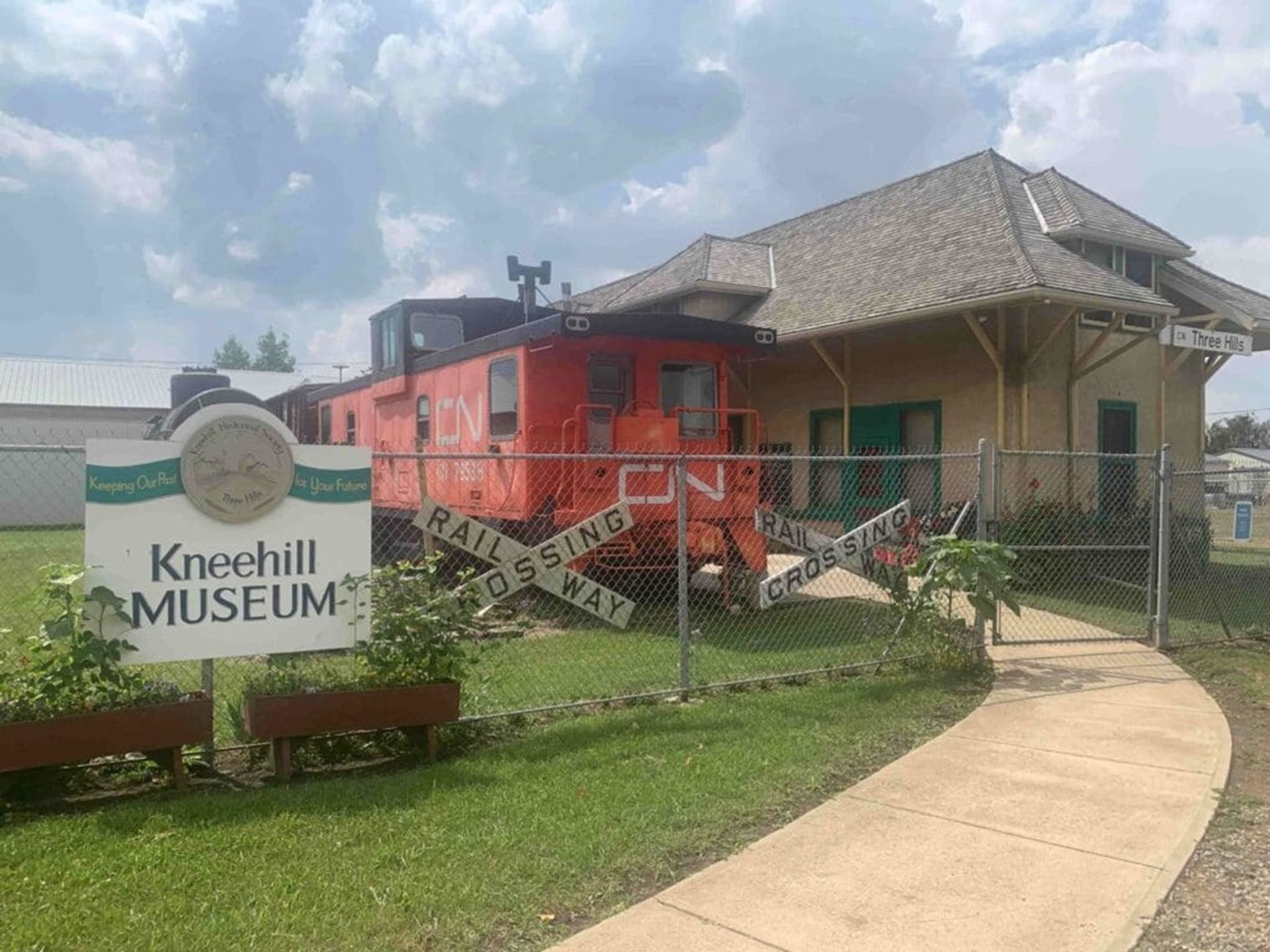 Kneehill Museum sign, orange CN rail car and historic station behind chain-link fence.