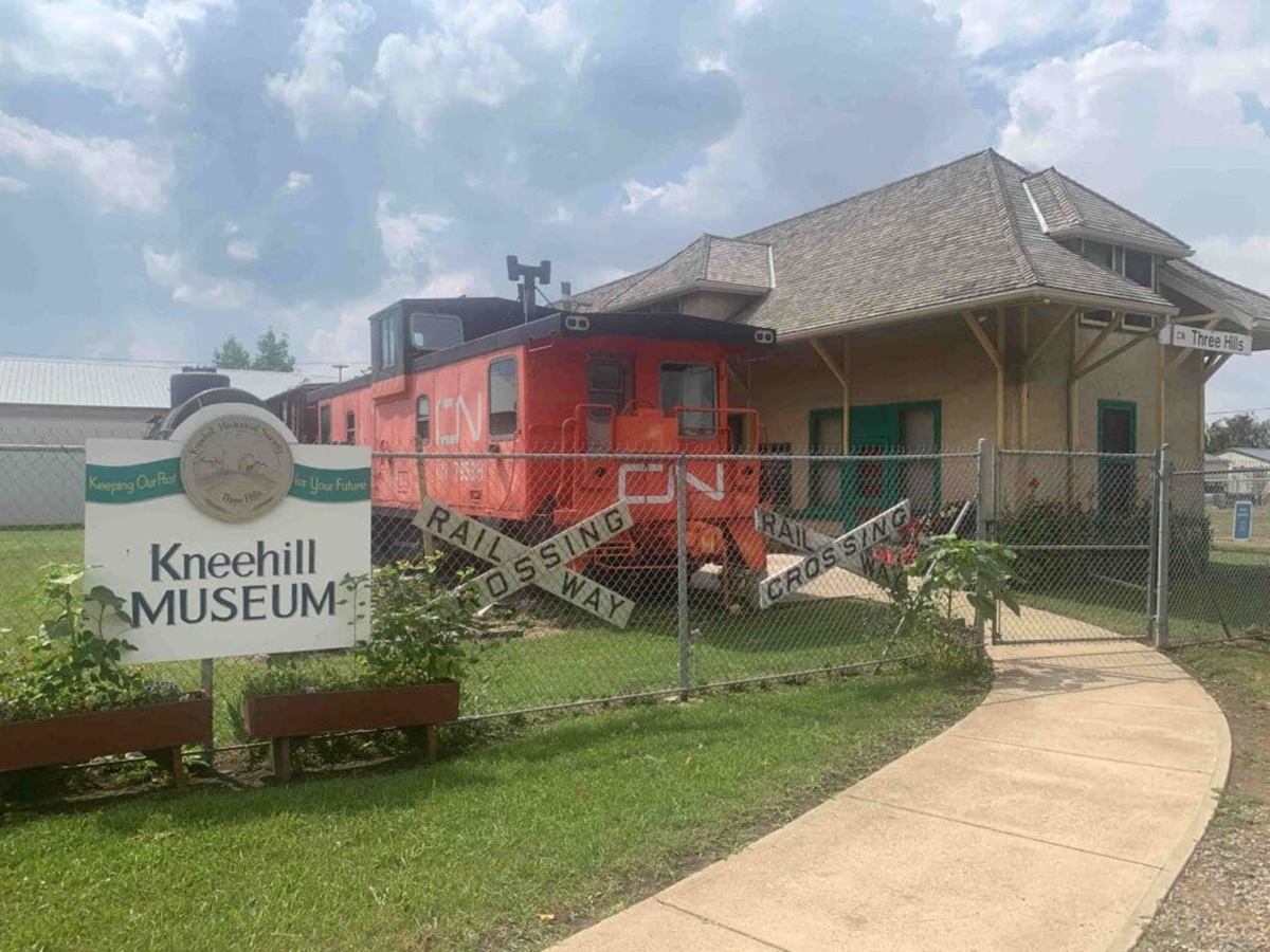 Kneehill Museum sign, orange CN rail car and historic station behind chain-link fence.