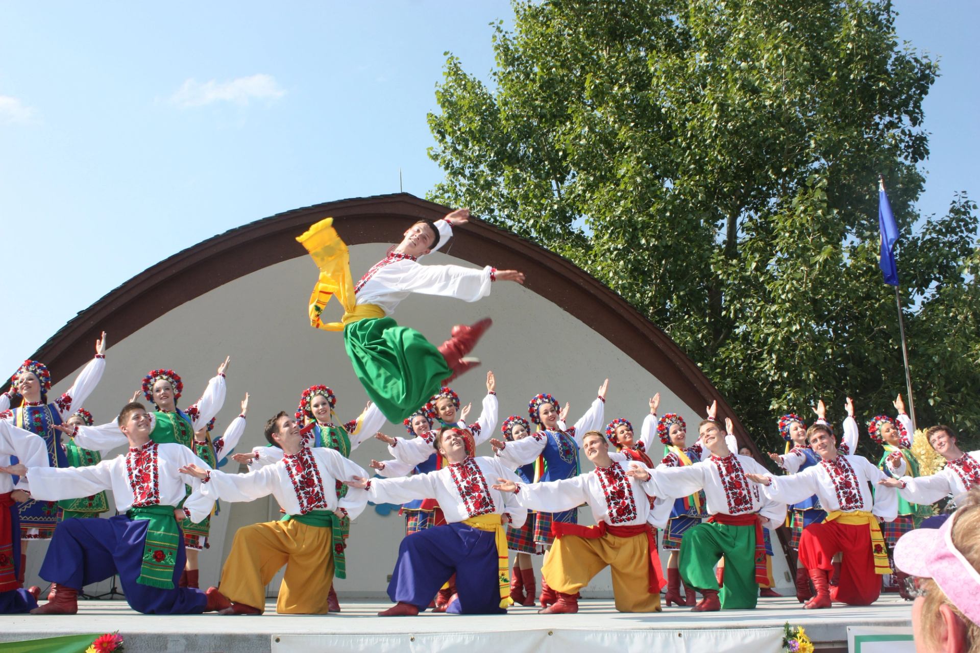 Ukrainian folk dancers perform on an outdoor stage, wearing colorful traditional costumes mid-leap.