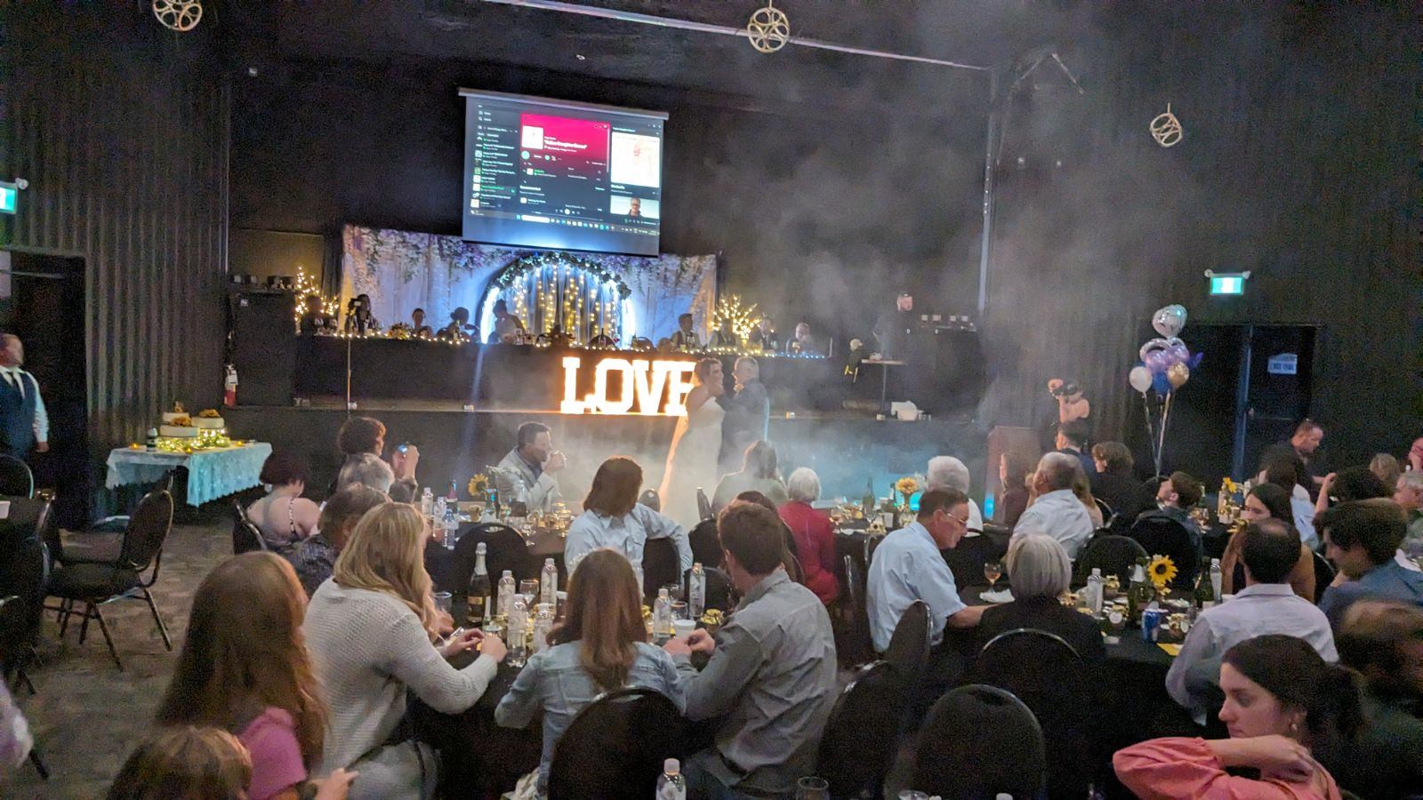 Wedding reception hall with couple dancing, LOVE sign, guests seated at decorated tables.