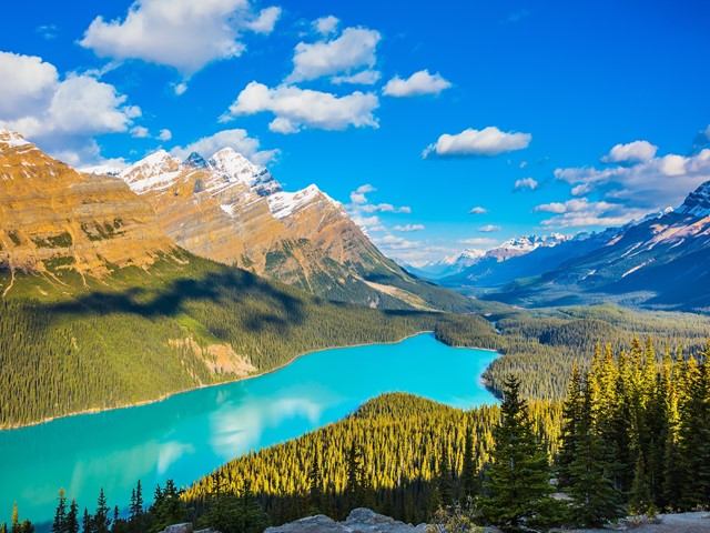 Turquoise mountain lake with pine forest and snowcapped peaks under blue sky with clouds.