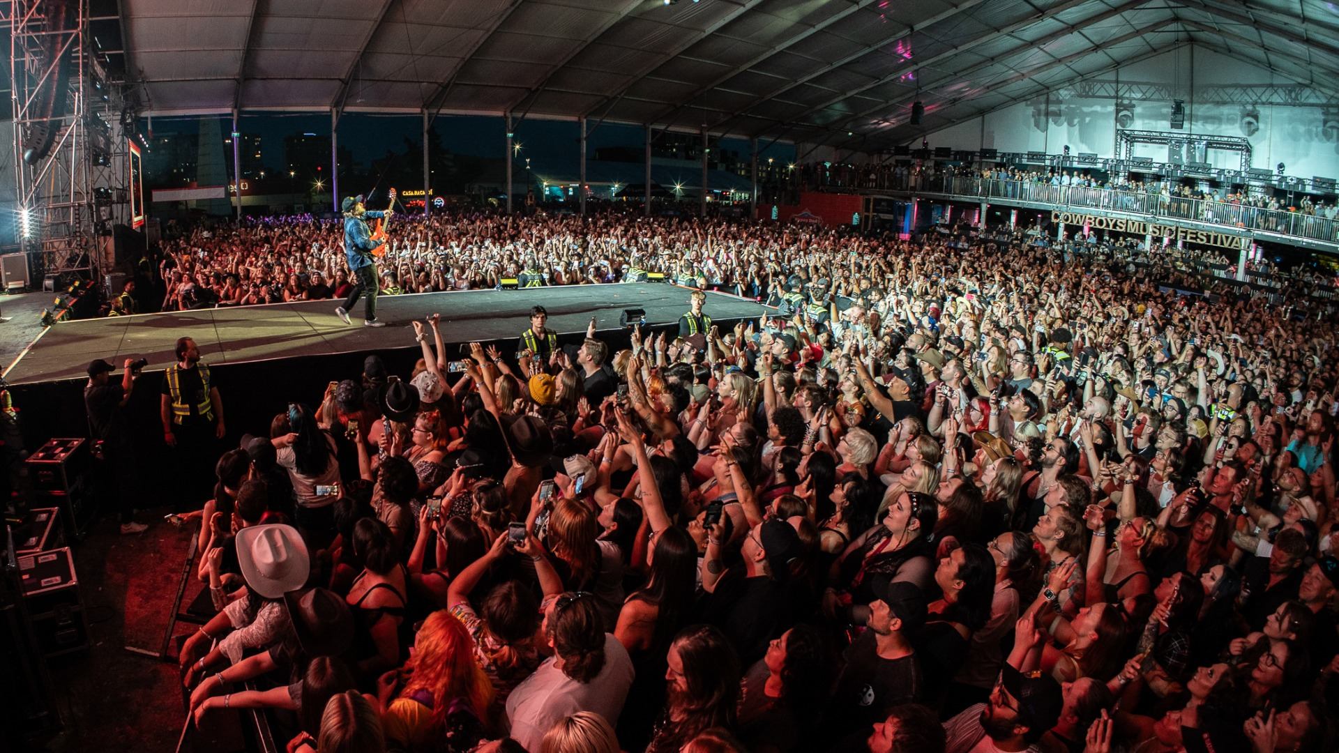 Large indoor concert crowd faces stage as guitarist performs under festival tent lighting.