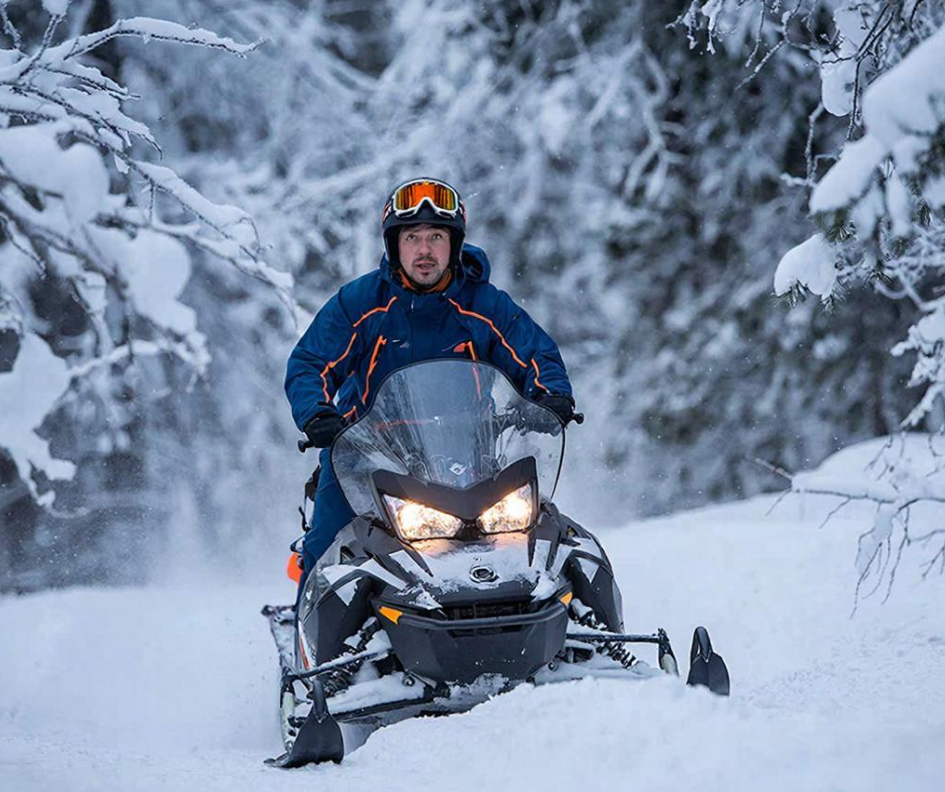 Snowmobiler rides along a snow-covered trail through dense forest.