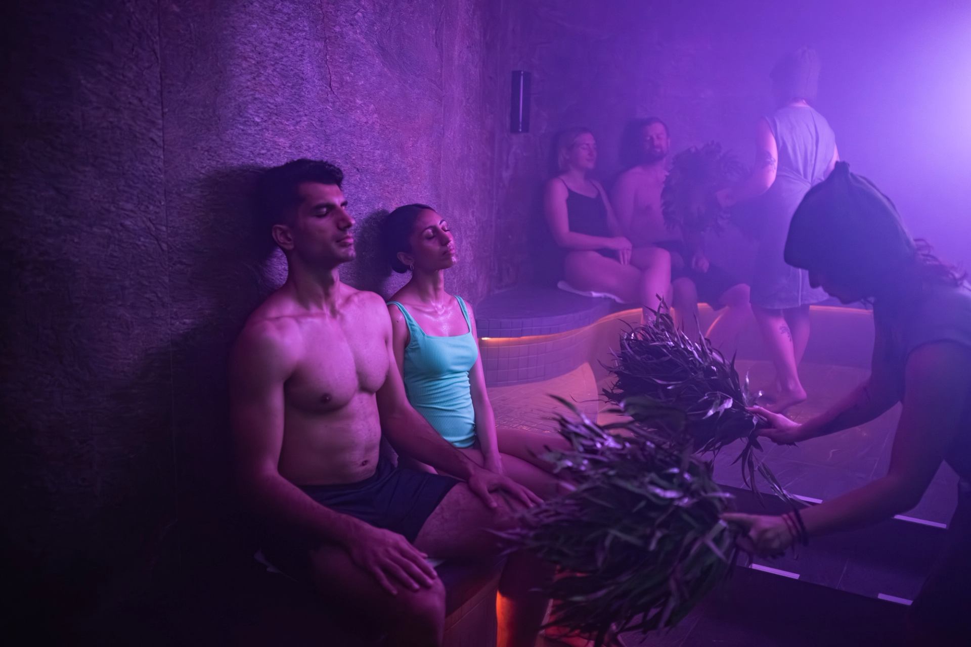 Guests seated in a steam room during a spa ritual, with soft purple lighting and fresh evergreen branches.