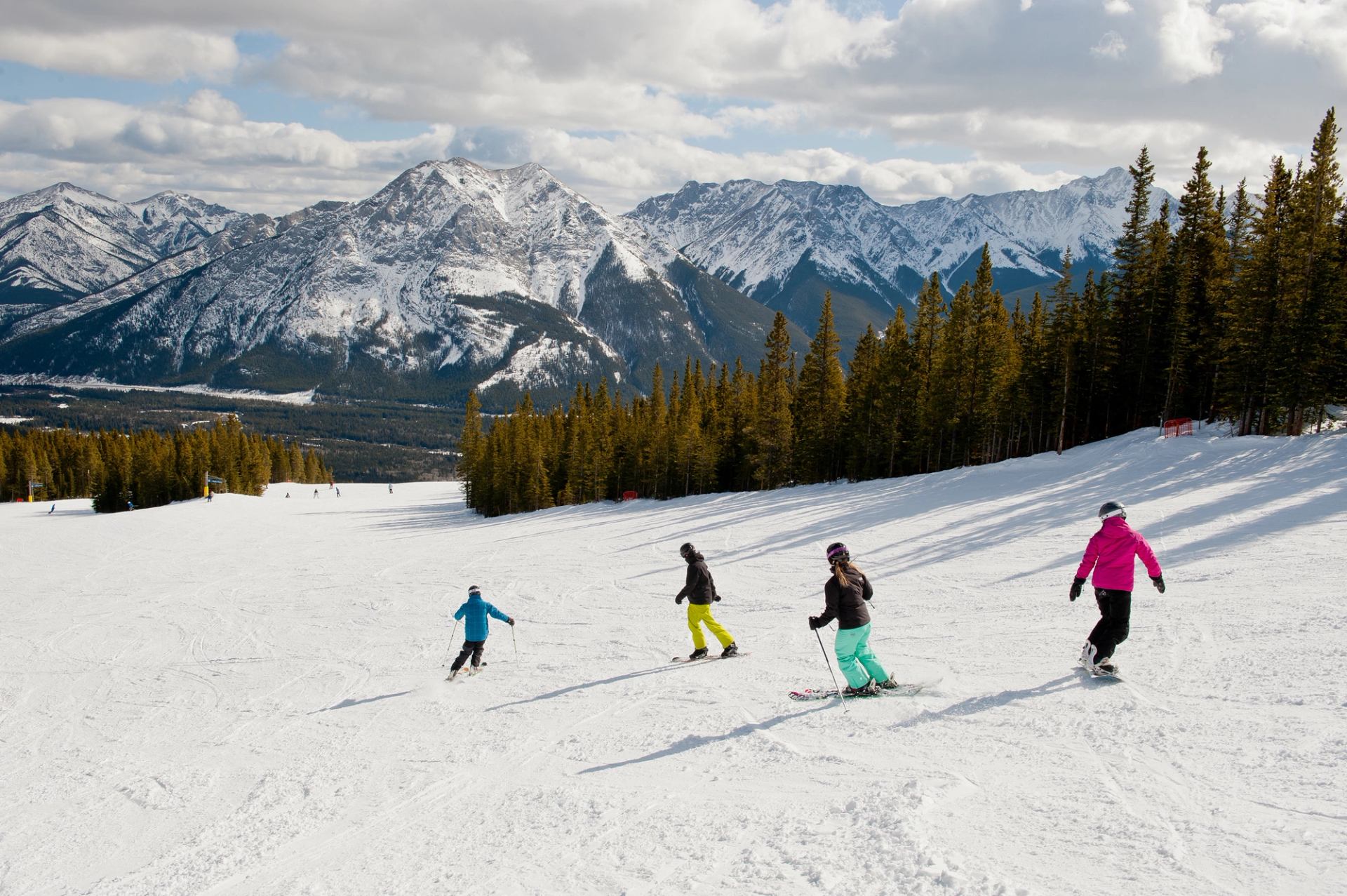 A group skiing down a snowy slope with tall peaks and forests in the background.