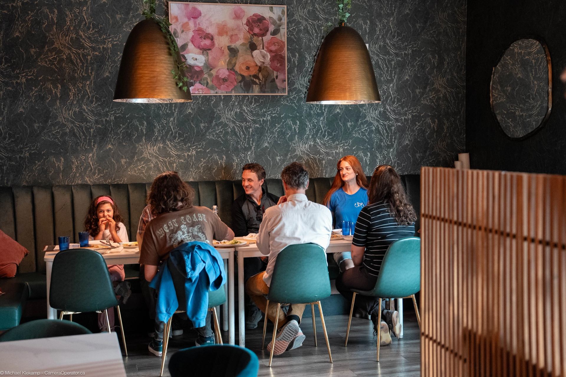 Group of people seated around a dining table in a modern restaurant with teal chairs and warm pendant lights.