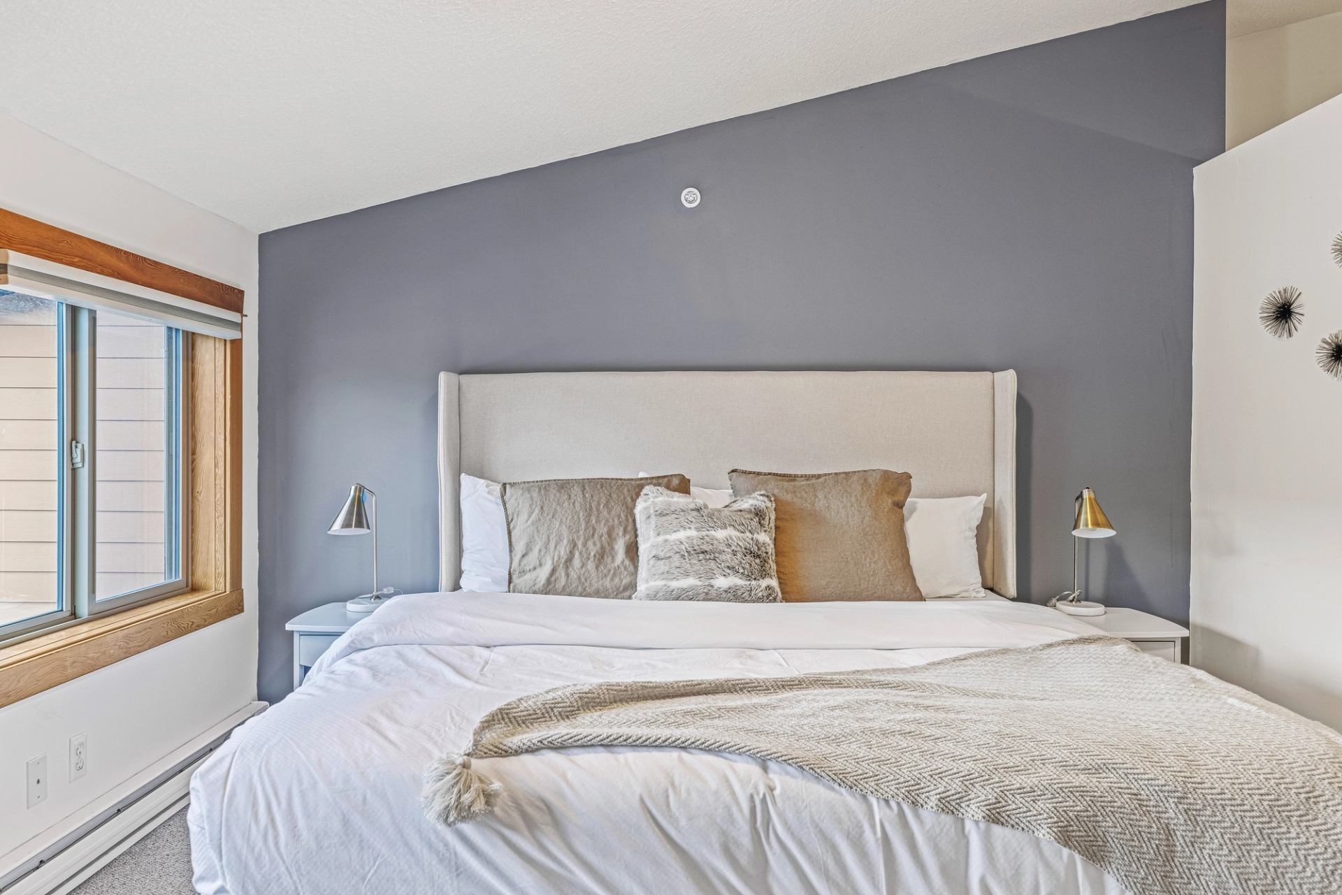 Bedroom with grey accent wall, large bed, neutral pillows and blanket, window and lamps.