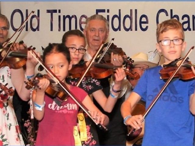 Group of people playing violins on stage under Old Time Fiddle Championship sign.