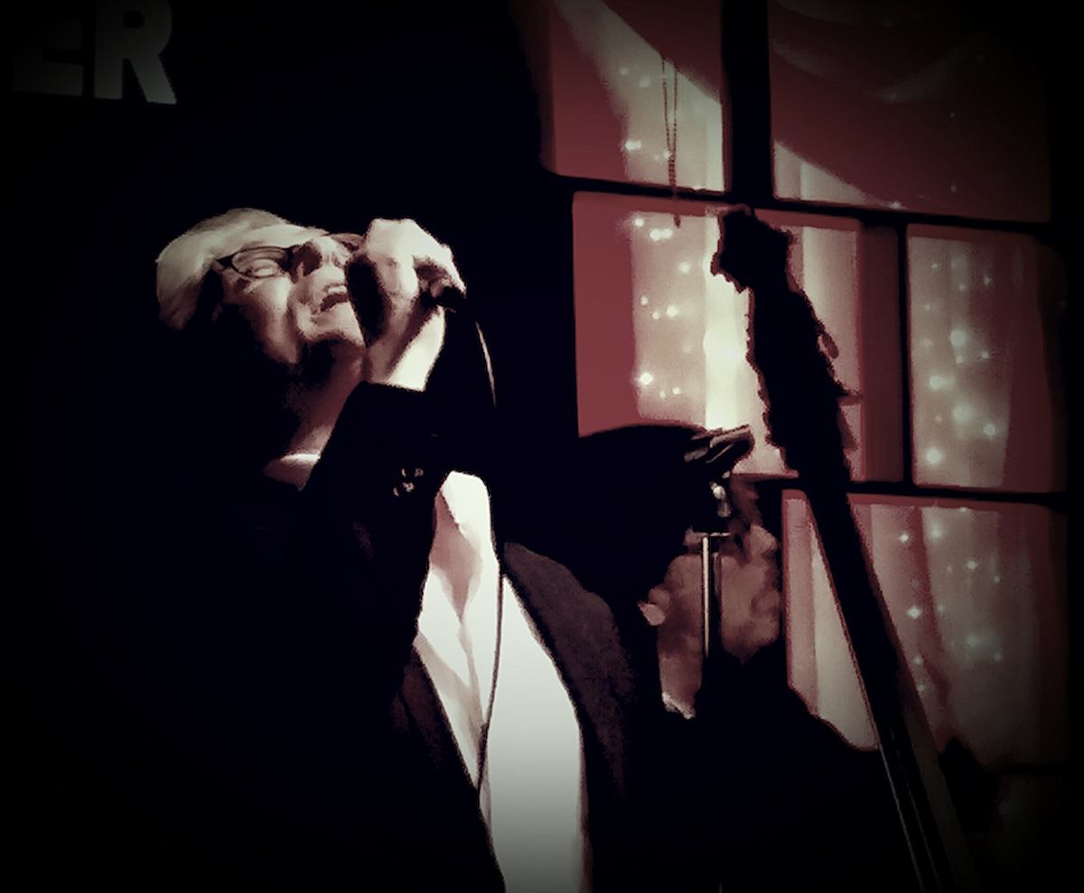 Performer singing into a microphone on stage, with lit backdrop and audience silhouettes.