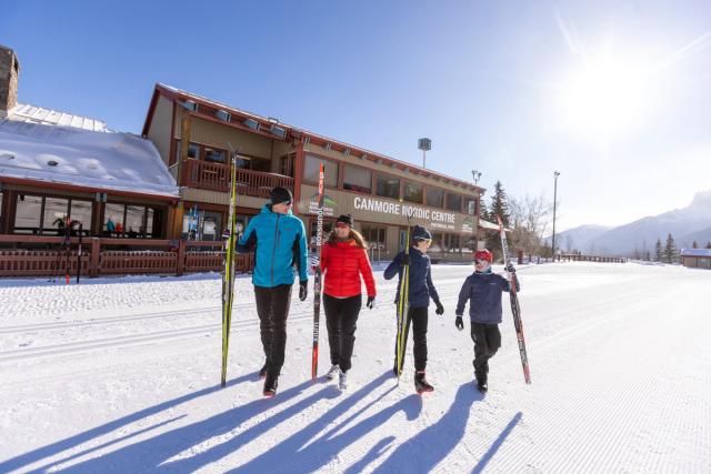 Family of four walking out of the ski lodge with their skis in hand in front of the Canmore Nordic Centre.