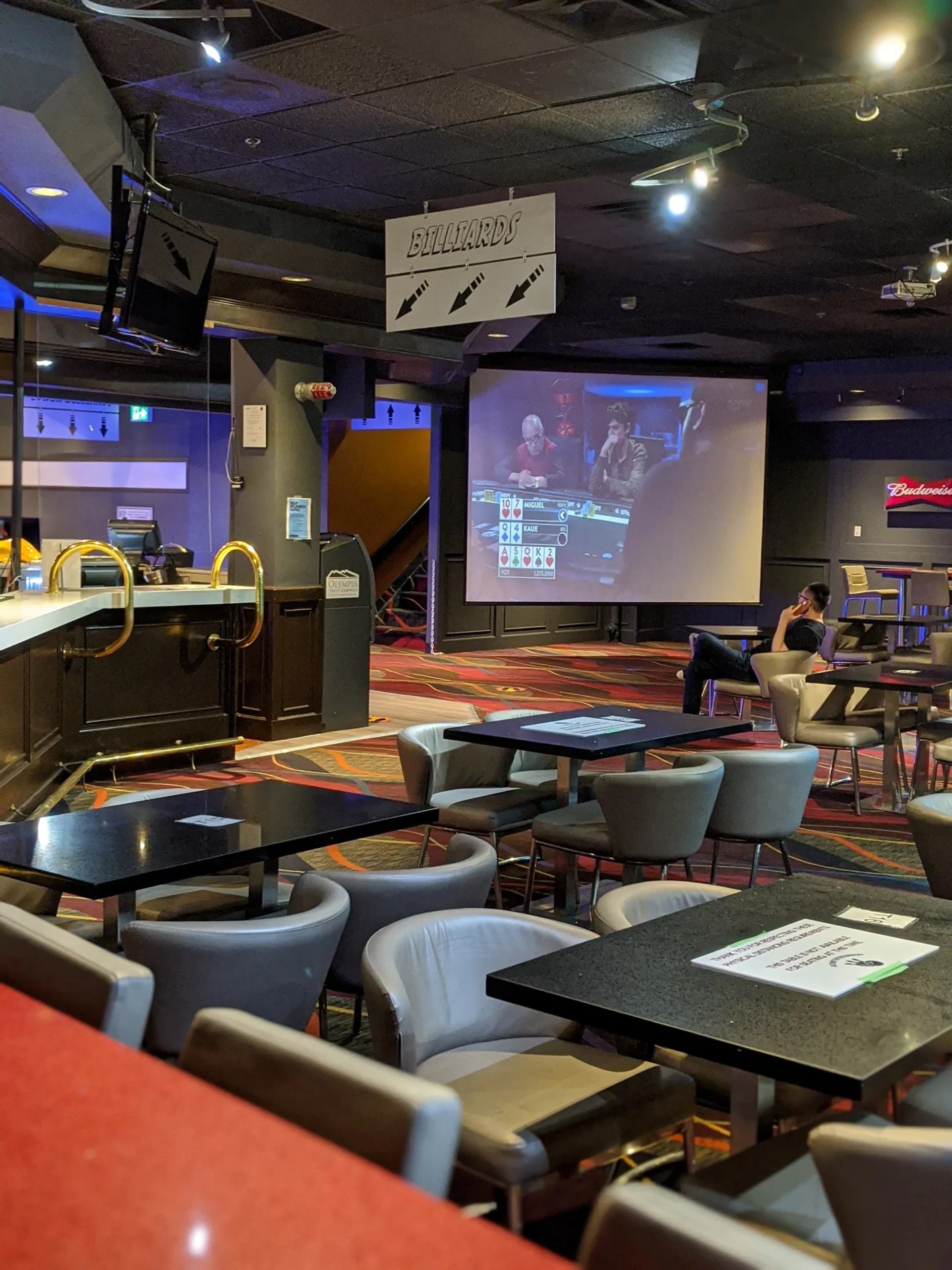 Sports bar interior with empty tables, billiards sign, and large screen showing poker game.