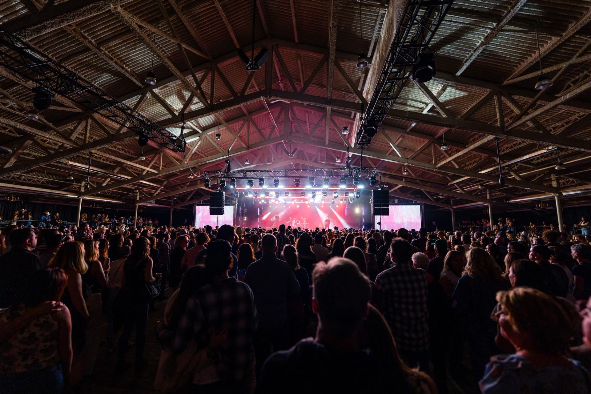 A large crowd views a band performing on a brightly lit stage with red lighting, under a high ceiling.