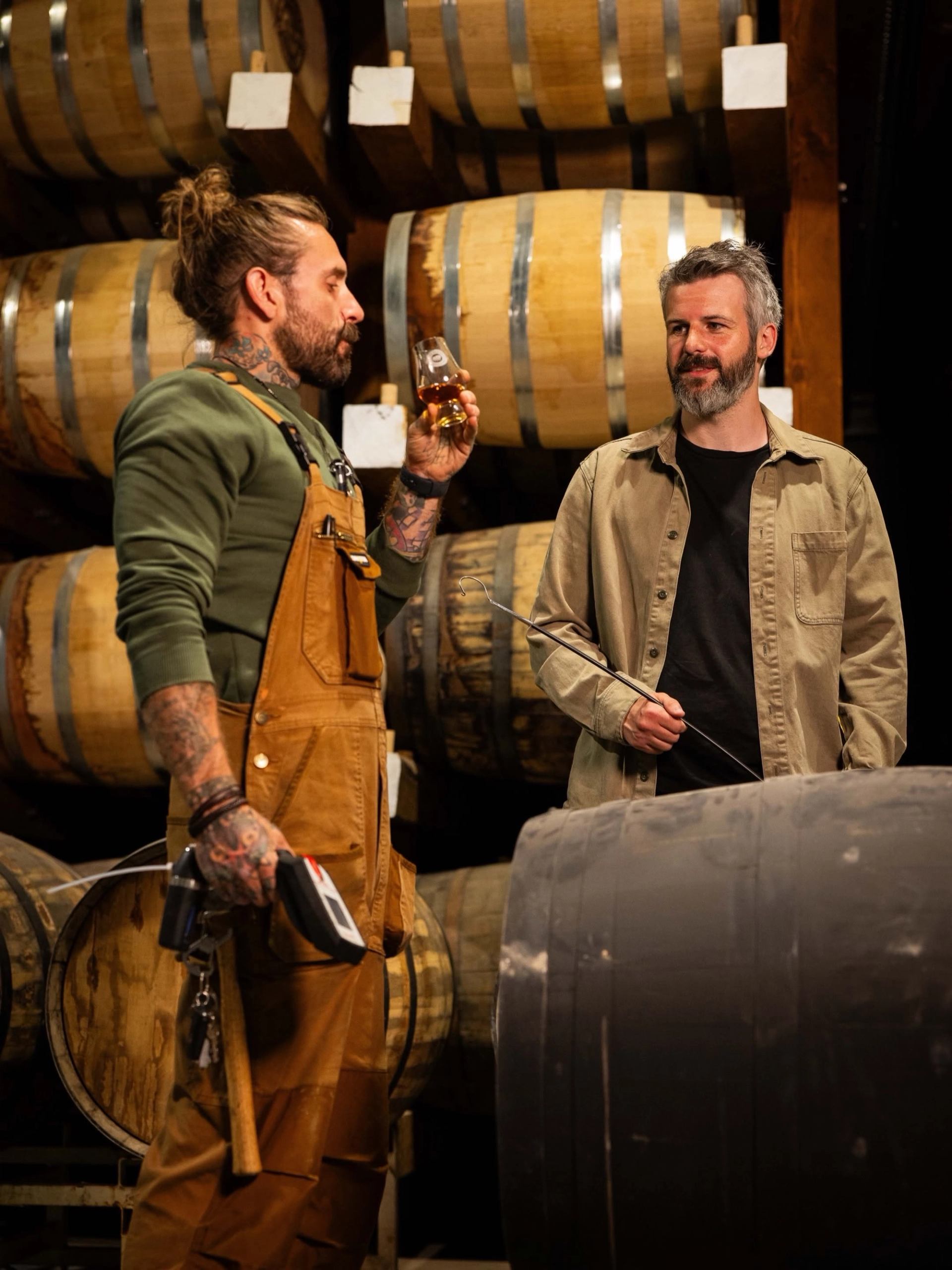 Two people stand among stacked barrels, discussing spirits and the aging process inside a room.