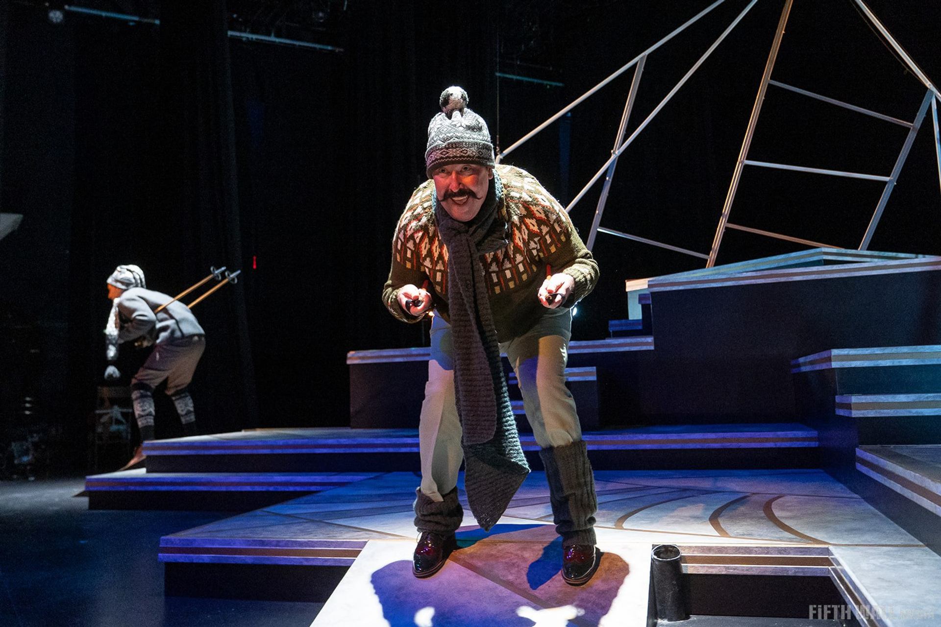 Stage performer in winter clothing leans forward under bright lights on geometric set.