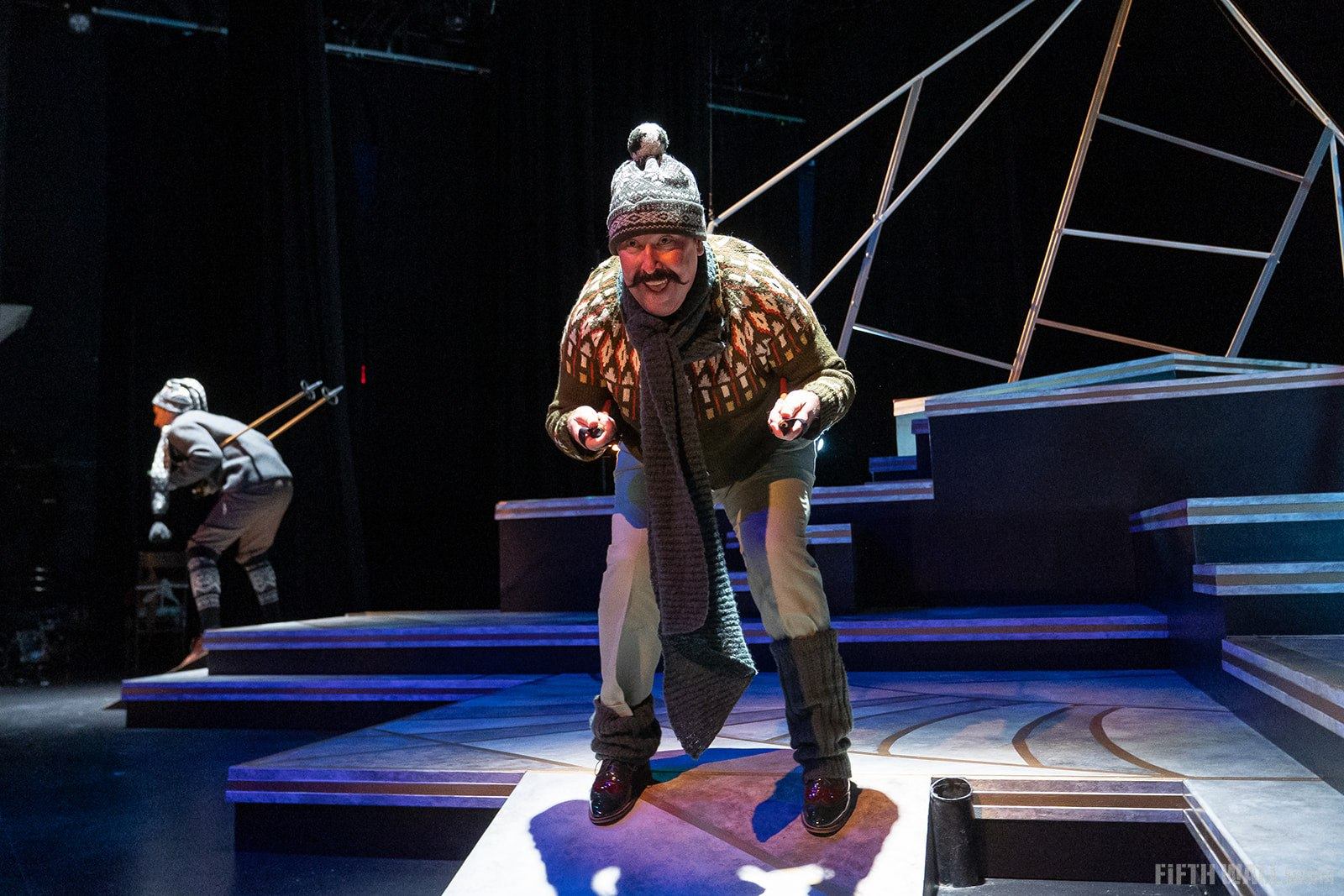 Stage performer in winter clothing leans forward under bright lights on geometric set.