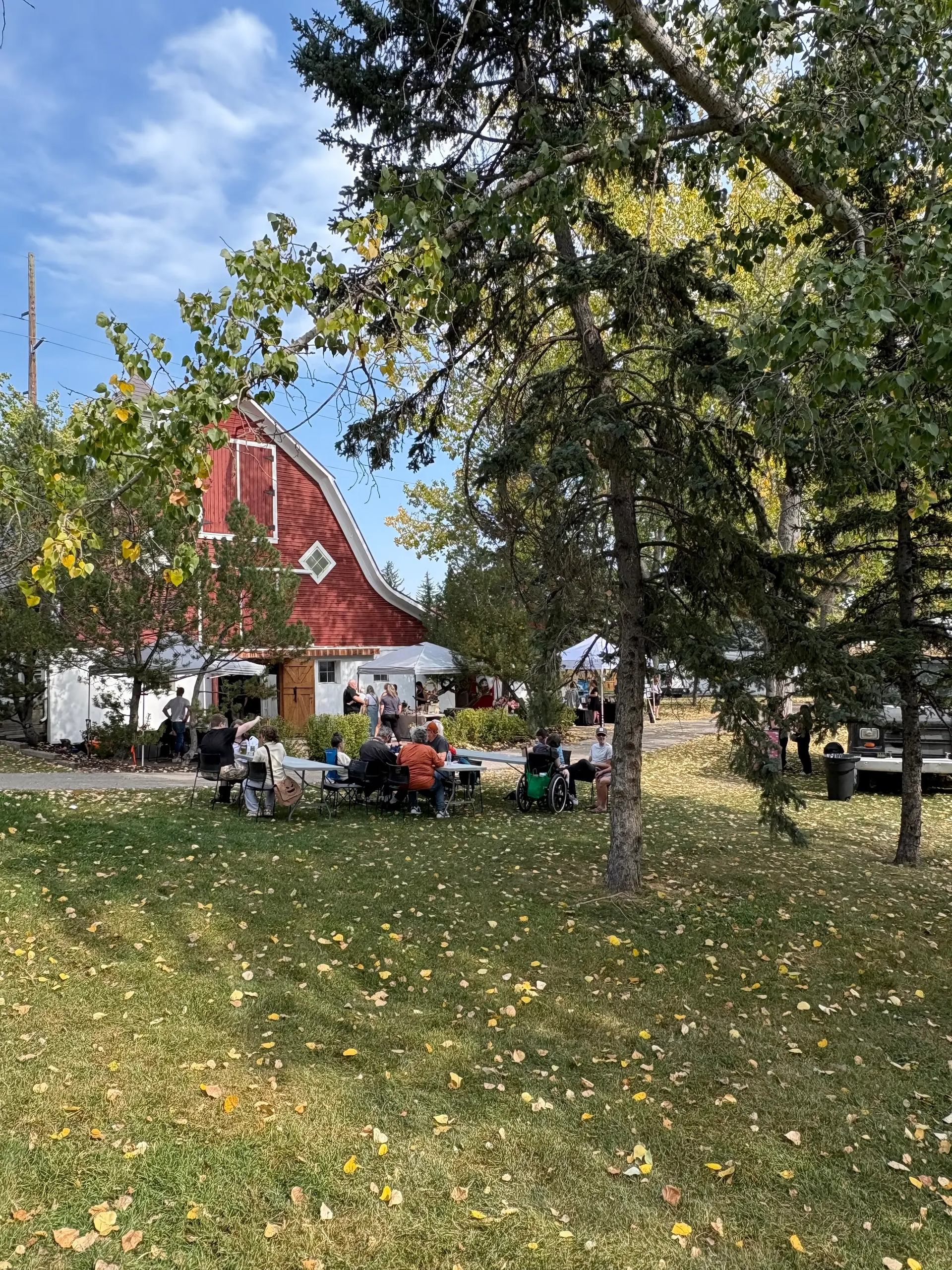 People sit at picnic tables on grass near red barn and white event tents.