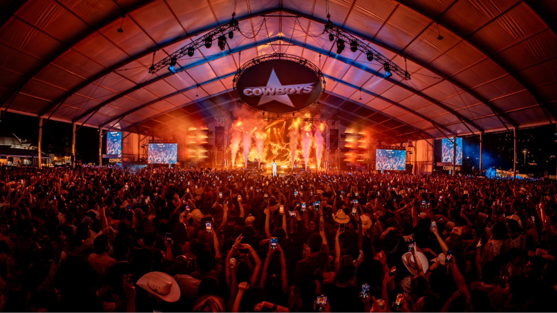 Crowd cheering as bright flames and lights illuminate the stage at Cowboys Music Festival.