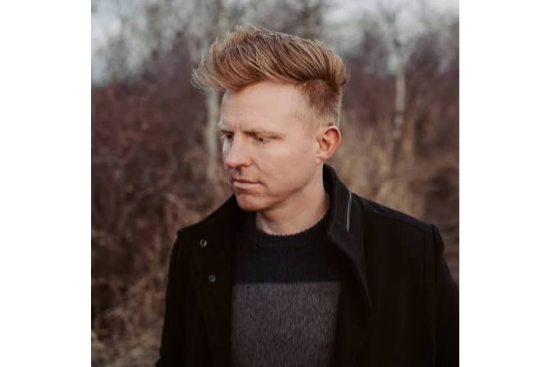 Person with styled blond hair and black jacket stands outdoors near leafless trees.